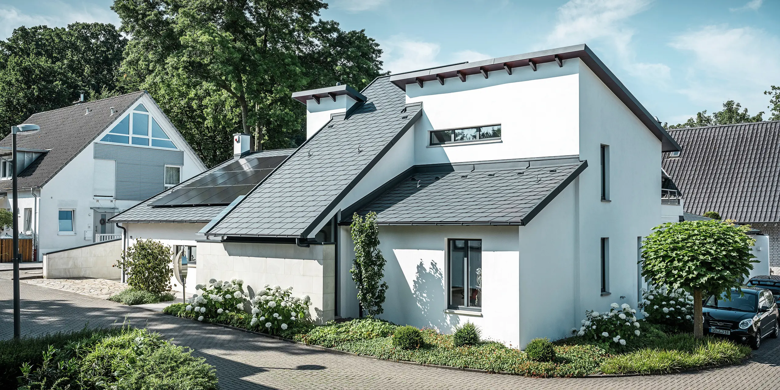 Modernt enfamiljshus med vit putsad fasad och mörkt PREFA-tak i aluminium. På taket syns integrerade solcellsmoduler och en takavvattning i aluminium i antracit. Byggnaden ligger i ett välskött bostadsområde med planterade trädgårdar, stenlagd uppfart och omgivande enfamiljshus.