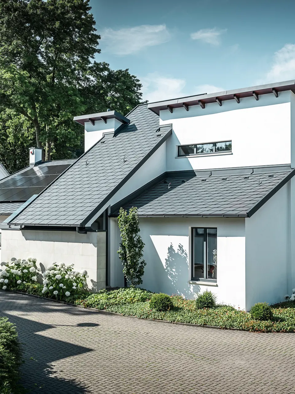 Modernt enfamiljshus med vit putsad fasad och mörkt PREFA-tak i aluminium. På taket syns integrerade solcellsmoduler och en takavvattning i aluminium i antracit. Byggnaden ligger i ett välskött bostadsområde med planterade trädgårdar, stenlagd uppfart och omgivande enfamiljshus.