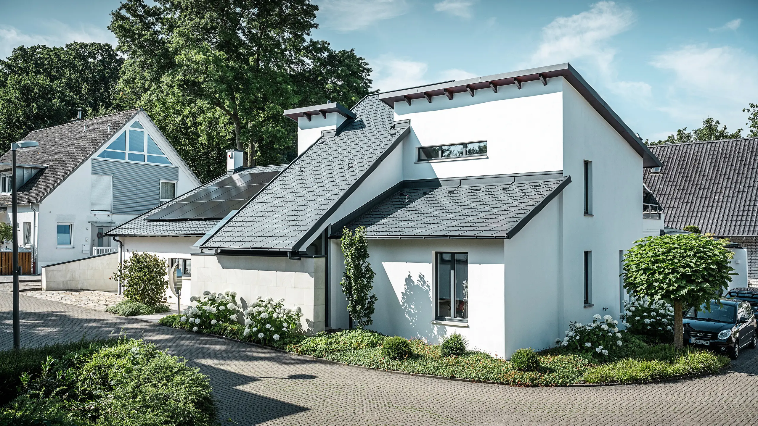 Modernt enfamiljshus med vit putsad fasad och mörkt PREFA-tak i aluminium. På taket syns integrerade solcellsmoduler och en takavvattning i aluminium i antracit. Byggnaden ligger i ett välskött bostadsområde med planterade trädgårdar, stenlagd uppfart och omgivande enfamiljshus.