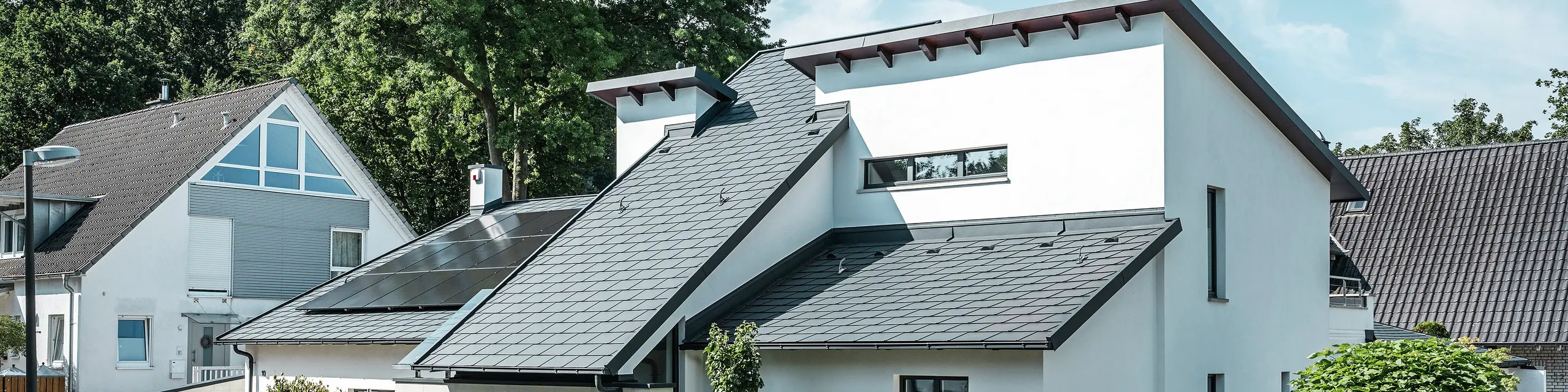 Modernt enfamiljshus med vit putsad fasad och mörkt PREFA-tak i aluminium. På taket syns integrerade solcellsmoduler och en takavvattning i aluminium i antracit. Byggnaden ligger i ett välskött bostadsområde med planterade trädgårdar, stenlagd uppfart och omgivande enfamiljshus.