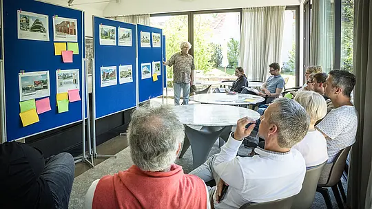 PREFARENZEN 2027 Expertendialog in einem hellen, modernen Seminarraum: Moderator Matthias Finkentey steht vor blauen Pinnwänden, auf denen Architekturprojekte mit PREFA Aluminiumlösungen präsentiert werden. Die Jury sitzt aufmerksam an runden Tischen, diskutiert und notiert Eindrücke. Große Fenster lassen Tageslicht herein, während draußen grüner Garten zu sehen ist. Die Szene zeigt die Verbindung von kreativer Architekturdiskussion, funktionaler Planung und Designbewertung. PREFA Aluminiumprodukte stehen für langlebige, ästhetische Dächer und Fassaden, die Innovation, Qualität und Inspiration in Architekturprojekten verkörpern.