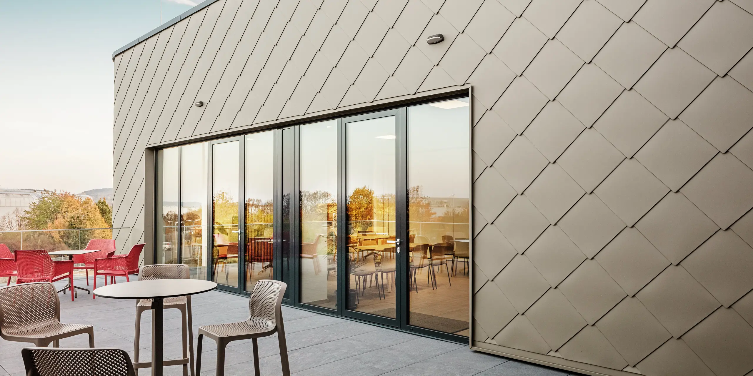 [Translate to Svenska:] Die Fassade des neuen PREFA Standortes in Törökbálint besticht durch die eleganten PREFA Wandrauten 44 × 44 in P.10 Prefabronze. Die präzise verlegte Aluminiumfassade sorgt für ein modernes, dynamisches Erscheinungsbild und bietet gleichzeitig maximalen Schutz vor Witterungseinflüssen. Die großzügige Glasfront verbindet Innen- und Außenbereich und schafft auf der großzügigen Terrasse eine helle und einladende Atmosphäre.