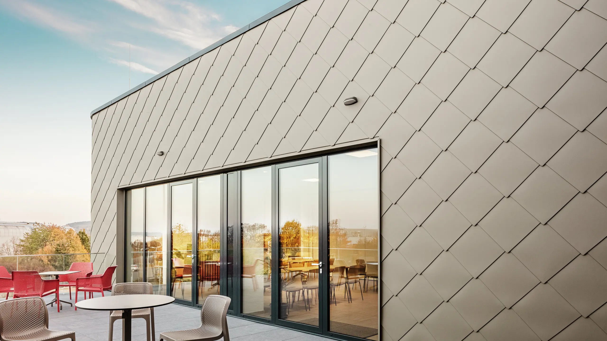 [Translate to Svenska:] Die Fassade des neuen PREFA Standortes in Törökbálint besticht durch die eleganten PREFA Wandrauten 44 × 44 in P.10 Prefabronze. Die präzise verlegte Aluminiumfassade sorgt für ein modernes, dynamisches Erscheinungsbild und bietet gleichzeitig maximalen Schutz vor Witterungseinflüssen. Die großzügige Glasfront verbindet Innen- und Außenbereich und schafft auf der großzügigen Terrasse eine helle und einladende Atmosphäre.