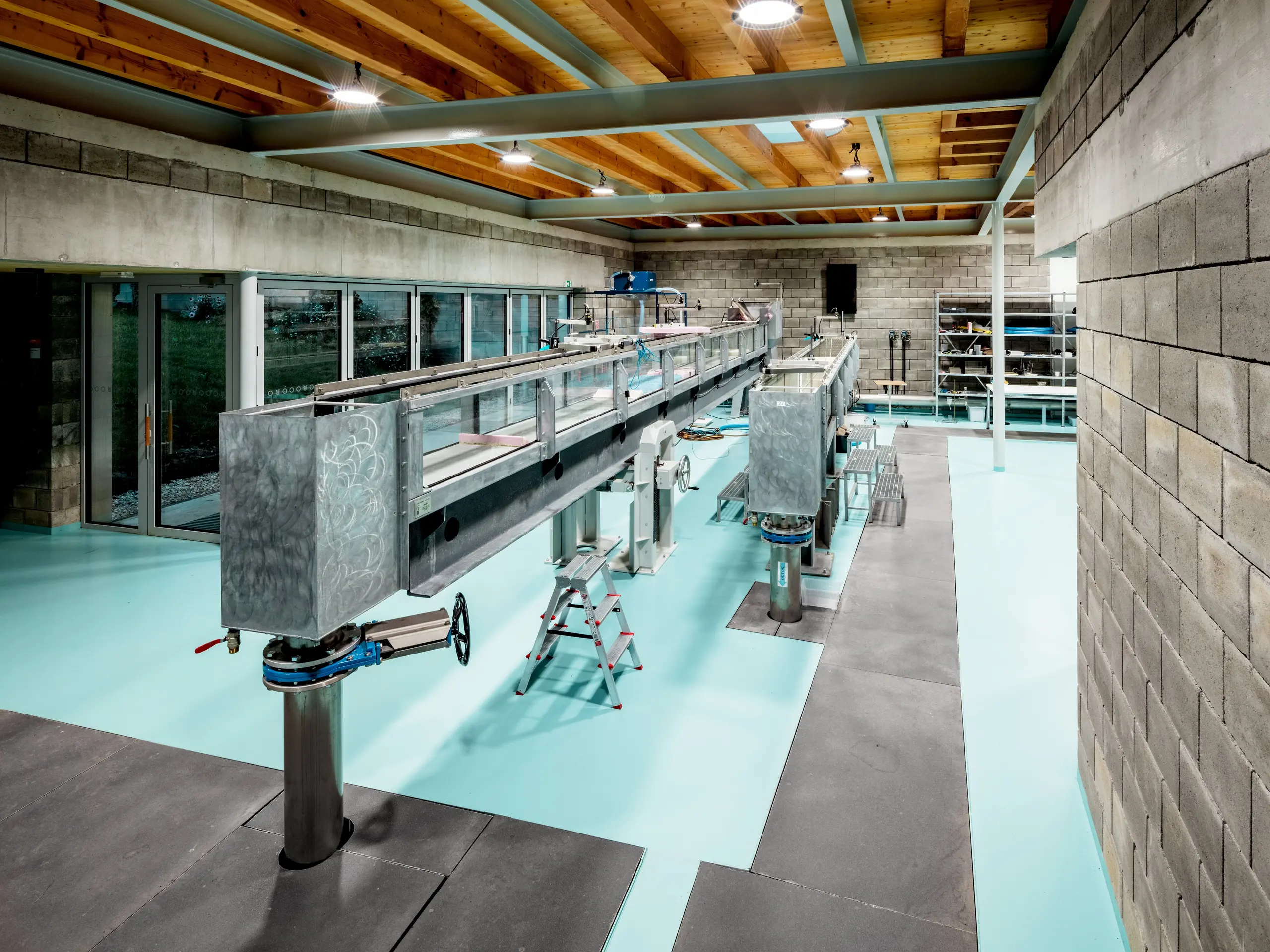 Moderné hydraulické laboratórium v Envirocentre Nitra zaujme dvoma dlhými prúdiacimi žľabmi určenými na vedecké experimenty zamerané na pohyb vody. Otvorenému výskumnému priestoru dominujú priznané oceľové nosníky, drevený strop v teplých odtieňoch, steny z pohľadových betónových tvárnic a tyrkysová podlaha. Veľkorysé presklené plochy vytvárajú svetlú a vzdušnú atmosféru a prepájajú inovatívny environmentálny výskum so súčasnou architektúrou a zachovanými prvkami pôvodnej stavby.