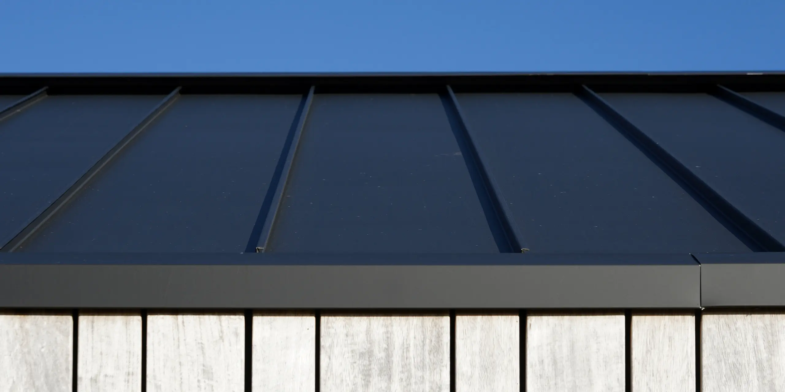 Detailaufnahme eines modernen Einfamilienhauses in Uithoorn, Niederlande, mit PREFALZ Winkelstehfalz-Eindeckung in Schwarzgrau. Das Bild zeigt die präzise Verarbeitung des hochwertigen Blechs von PREFA, kombiniert mit natürlichen Holzelementen an der Fassade. Die klare Linienführung des robusten Aluminiumdachs unterstreicht die langlebige und wetterbeständige Bauweise.
