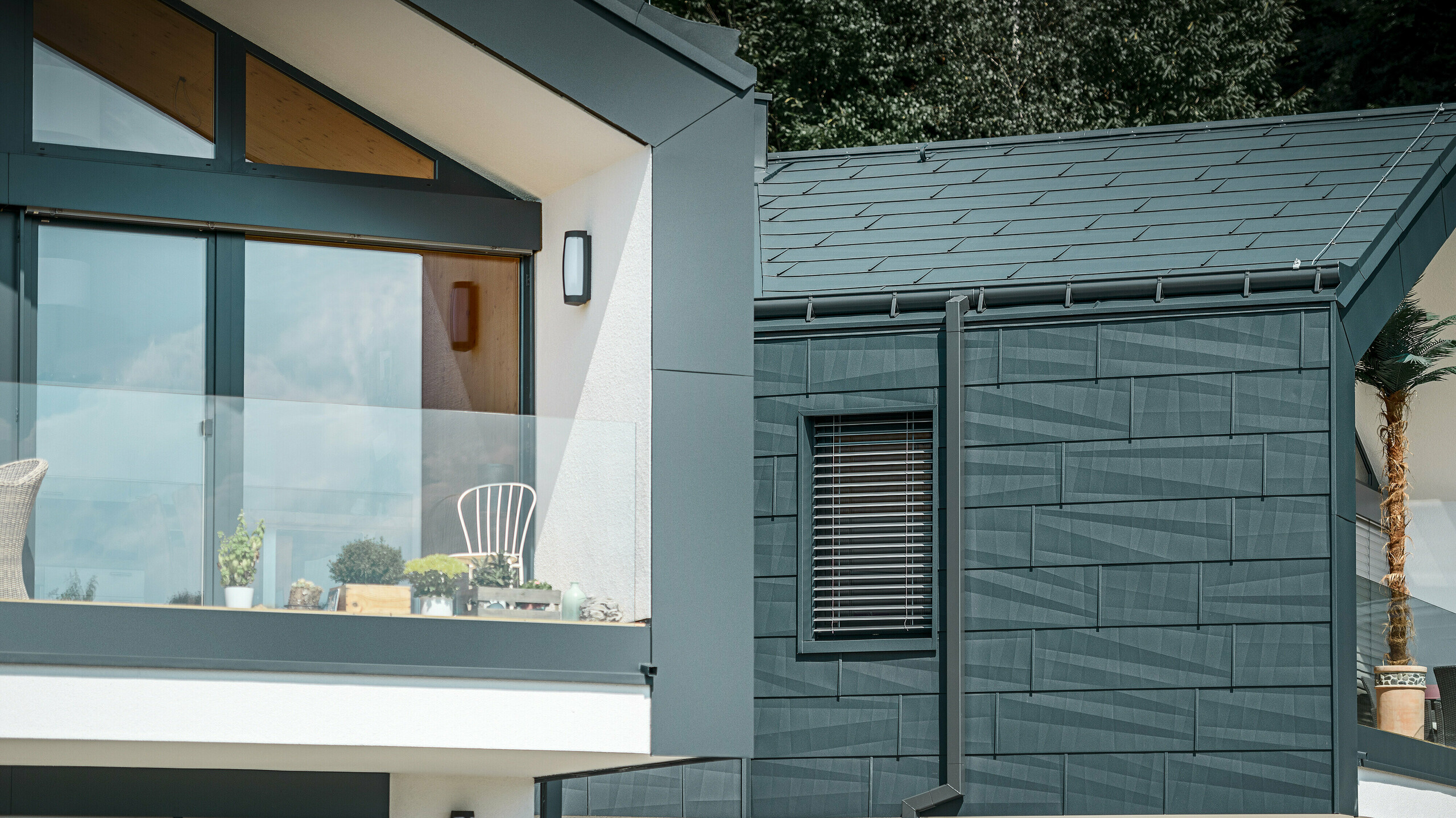 Bilden visar ett modernt parhus med en stilren fasad i aluminium. Fasaden är i en mörk antracitfärg och har en strukturerad yta tack vare PREFA tak- och fasadpaneler FX.12. På taket har en takränna och ett fyrkantigt avloppsrör från PREFA monterats. En balkong med glasräcke syns på övervåningen, medan fönster med lamelljalusier finns på nedervåningen. Arkitekturen är klar och minimalistisk, och i bakgrunden syns träd som skapar en naturlig atmosfär.