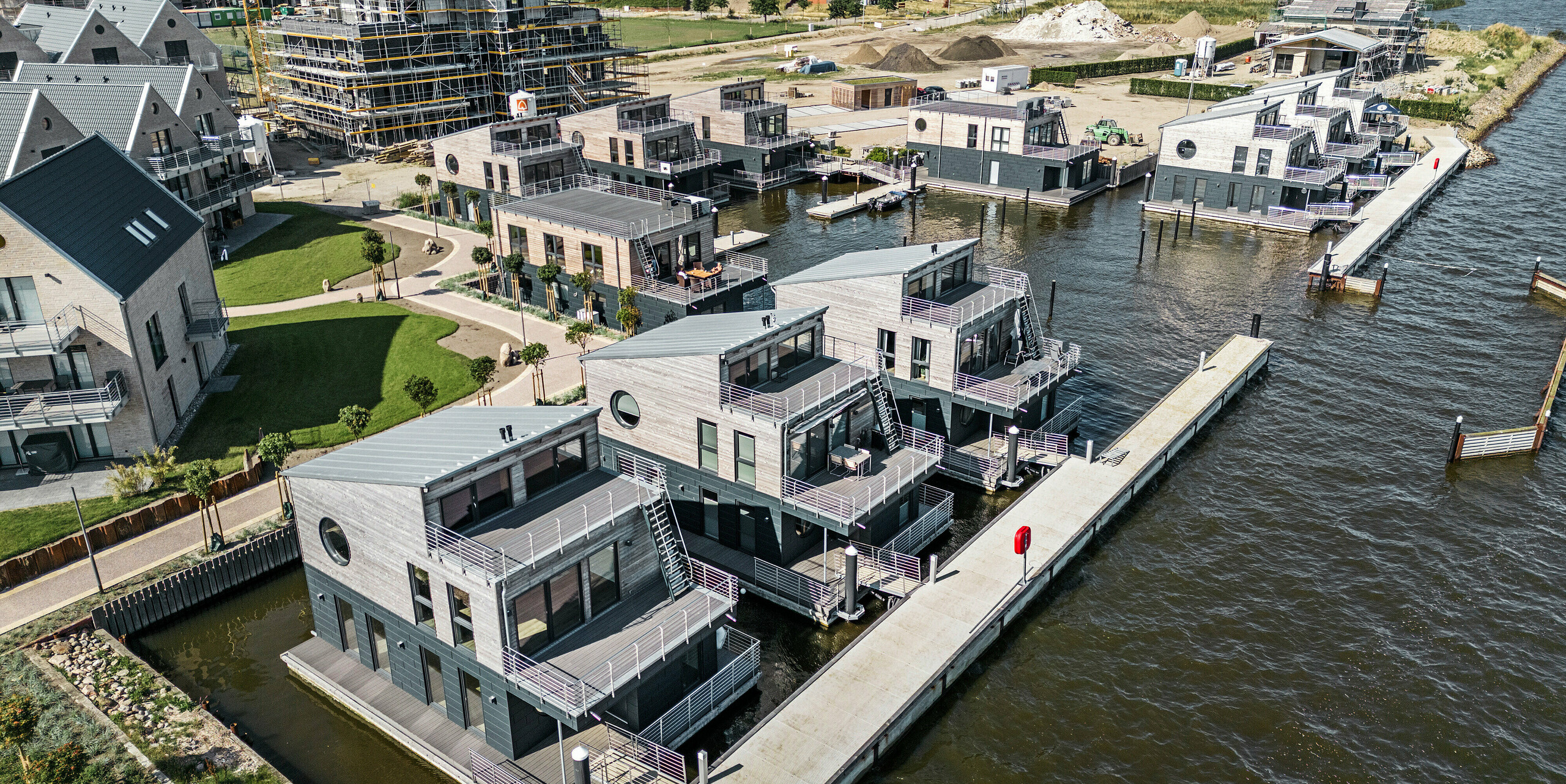 Luftaufnahme der innovativen Wasserhaus-Siedlung im Pionierhafen Schleswig, eingebettet in einer maritimen Umgebung und angrenzender Bebauung im Aufbau. Die elf schwimmenden Domizile, charakteristisch verkleidet mit PREFA Siding.X in P.10 Anthrazit, harmonieren mit den Holzelementen an der Fassade. Die PREFA Aluminiumprodukte stehen für ihre Beständigkeit gegenüber der direkten Wassernähe. Die architektonische Symbiose von funktionaler Eleganz und natürlicher Ästhetik entlang des Ufers unterstreicht die Exklusivität dieser Wohnanlage, die primär als Feriendestination dient.