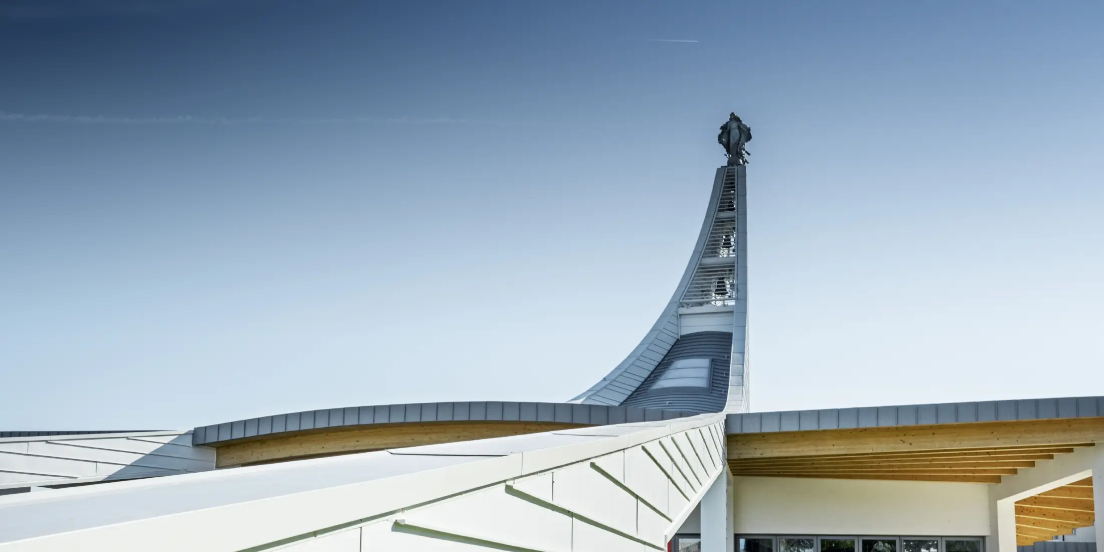 Modernes Kirchengebäude mit geschwungener Dachkonstruktion aus hellen Aluminiumpaneelen. Im Hintergrund erhebt sich ein hoher, elegant gebogener Turm mit einer Statue an der Spitze. Die Architektur kombiniert klare Linien mit offenen Glasflächen und Holzelementen unter dem Dach. Der Himmel ist wolkenlos und betont die helle, futuristische Gestaltung des Bauwerks