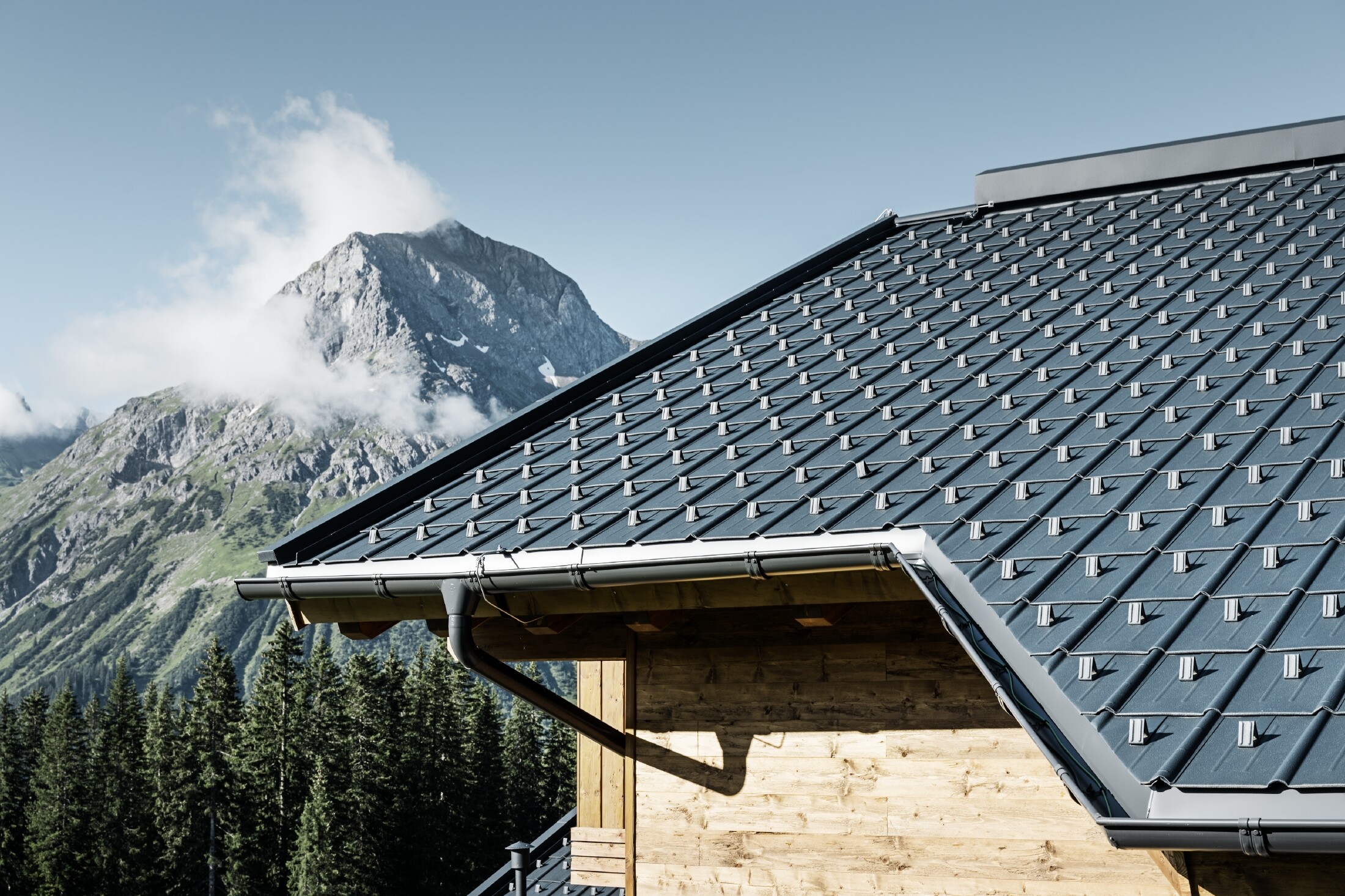 PREFA takplatta i alpin insats – Takplatta i antracit lagt på fjällstuga i Lech am Arlberg med berg i bakgrunden. PREFA takränna i antracit; träfasad