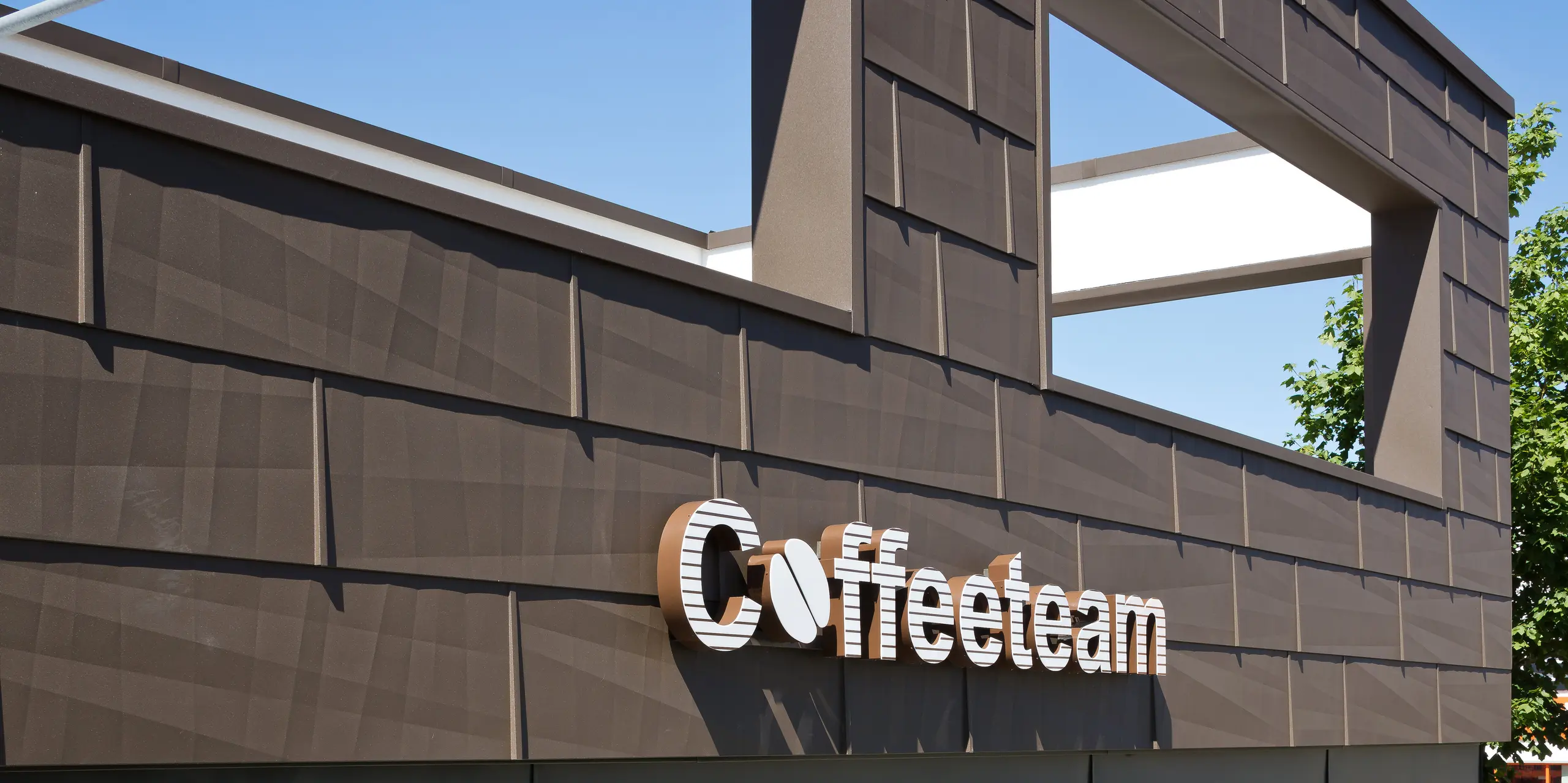 Detailansicht des Coffeeteam-Firmenschilds an der Fassade der Unternehmenszentrale in München. Die Fassade wurde mit PREFA Fassadenpaneelen FX.12 in der Farbe P.10 Braun verkleidet. Die außergewöhnliche Oberflächenstruktur der Aluminiumfassade erzeugt durch Licht und Schatten eine lebendige, dreidimensionale Wirkung. Das markante Firmenlogo hebt sich wirkungsvoll von der modernen Gebäudehülle ab. Die langlebige, witterungsbeständige und nahezu wartungsfreie PREFA-Aluminiumfassade verbindet anspruchsvolles Design mit nachhaltiger Funktionalität.