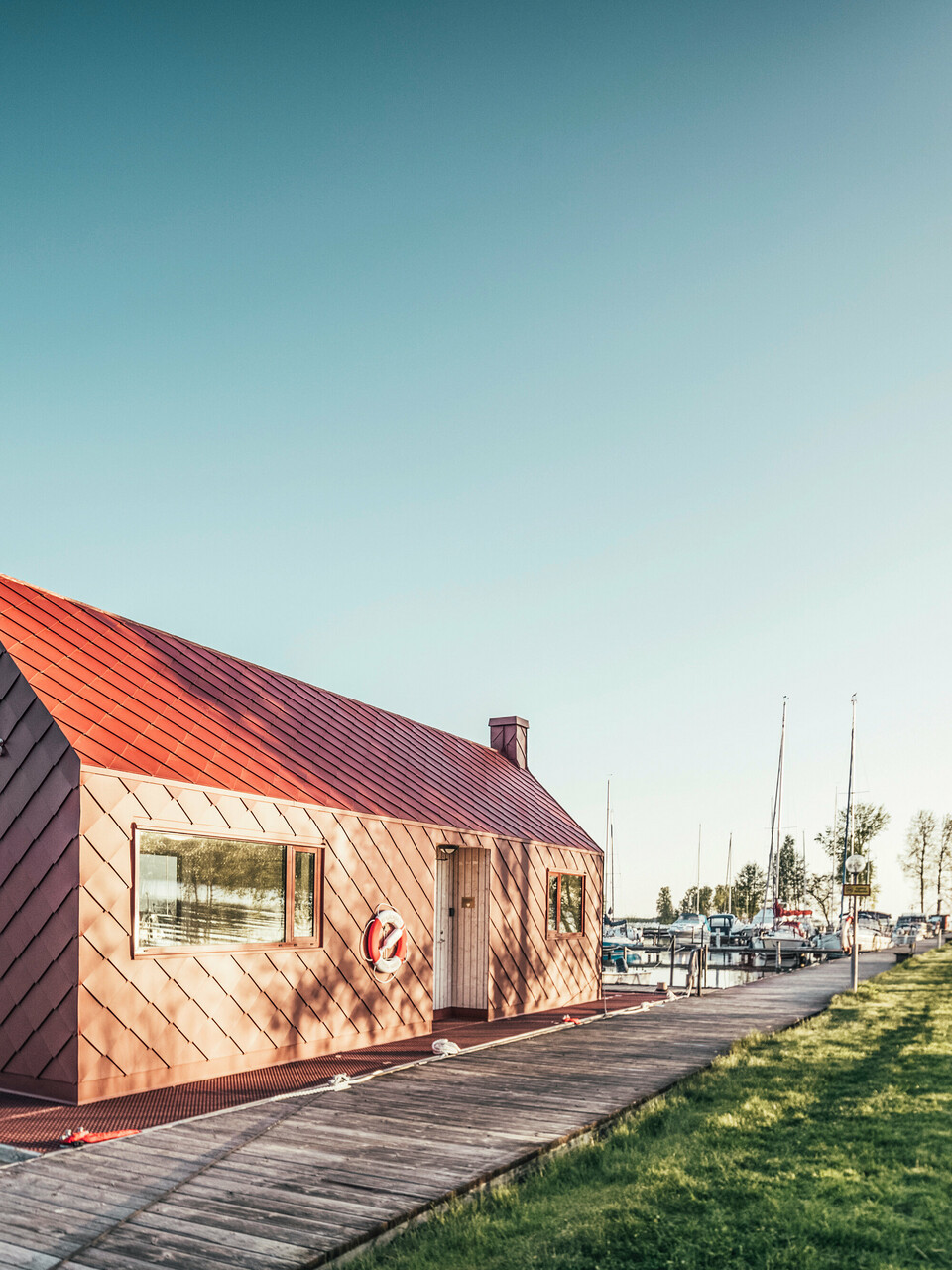 Seenotrettungsstation in harmonischer Umgebung am Hjälmaren See in Schweden, mit einem oxydroten PREFA Dach und Fassadenruten 29×29. Die Station liegt am Ende eines Holzstegs, der in einen Yachthafen führt, wo Boote sanft auf dem Wasser schaukeln. Grüne Bäume rahmen das Bild, das die ruhige, aber bereitwillige Atmosphäre einer Rettungsstation bei goldenem Morgenlicht einfängt.
