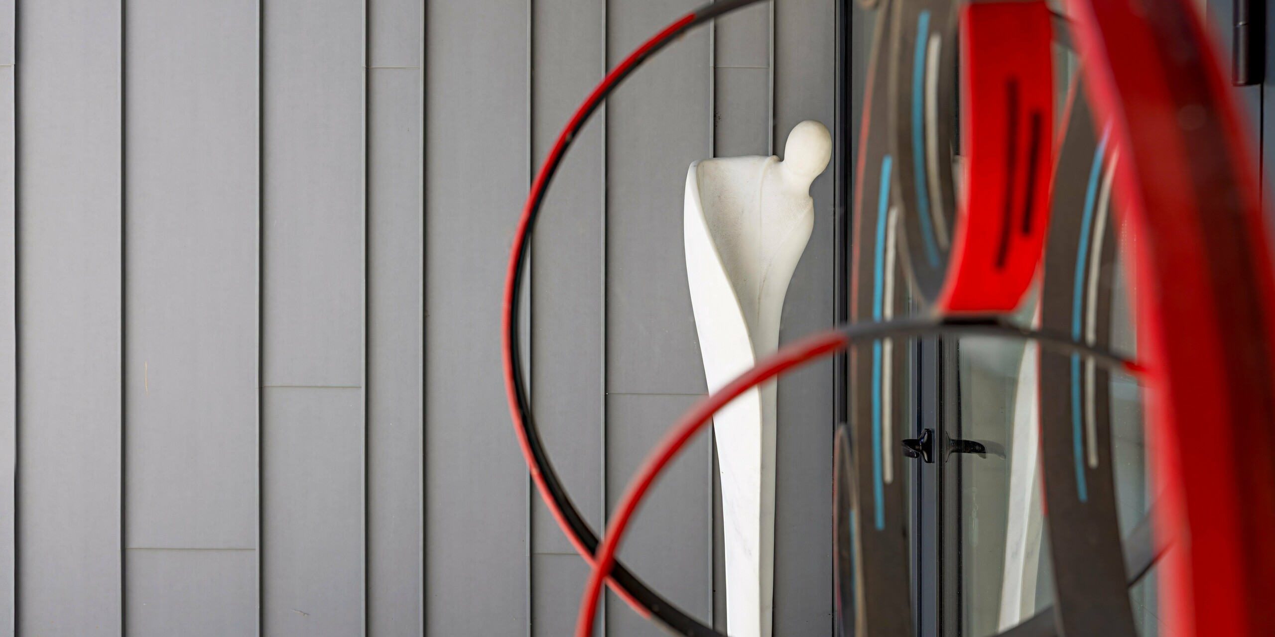 Eine abstrakte Kunstskulptur aus geschwungenen roten und schwarzen Ringen rahmt eine weiße Menschenfigur ein und tritt in einen faszinierenden Dialog mit der PREFA Aluminiumfassade in Patinagrau der Scuola Arte e Mestieri F. Bertazzoni in Suzzara. Die präzise vertikal gegliederten PREFALZ Fassadenelemente bilden eine ebenso ruhige wie edle Kulisse, die die künstlerische Installation optimal zur Geltung bringt. Die samtig matte Oberfläche der PREFA Aluminiumverkleidung unterstreicht die hochwertige Qualität und zeitlose Beständigkeit des gesamten Gebäudeensembles.