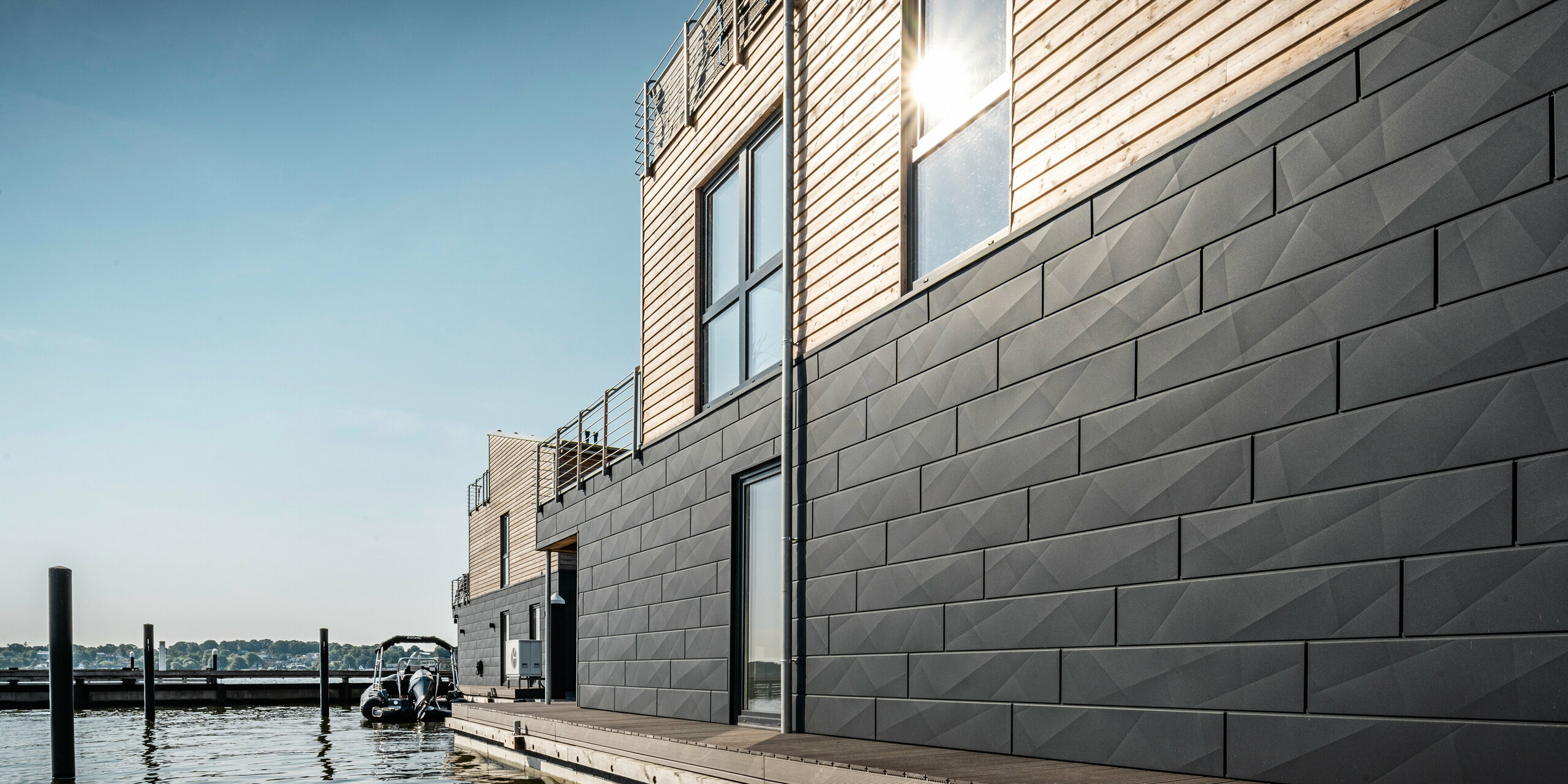 Moderne Wasserhäuser am Ufer der Schlei mit einer robusten und nachhaltigen Außenhülle aus Holz und Aluminium. Die erste Etage der schwimmenden Häuser im Pionierhafen Schleswig ist umhüllt von PREFA Siding.X in P.10 Anthrazit, einem widerstandsfähigen und rostsicheren Aluminium-Fassadensystem, das Feuchtigkeit effizient durch eine vorgehängt hinterlüftete Fassaden-Konstruktion abführt. Zudem bietet die Aluminiumfassade eine markante Optik im Knitterlook.