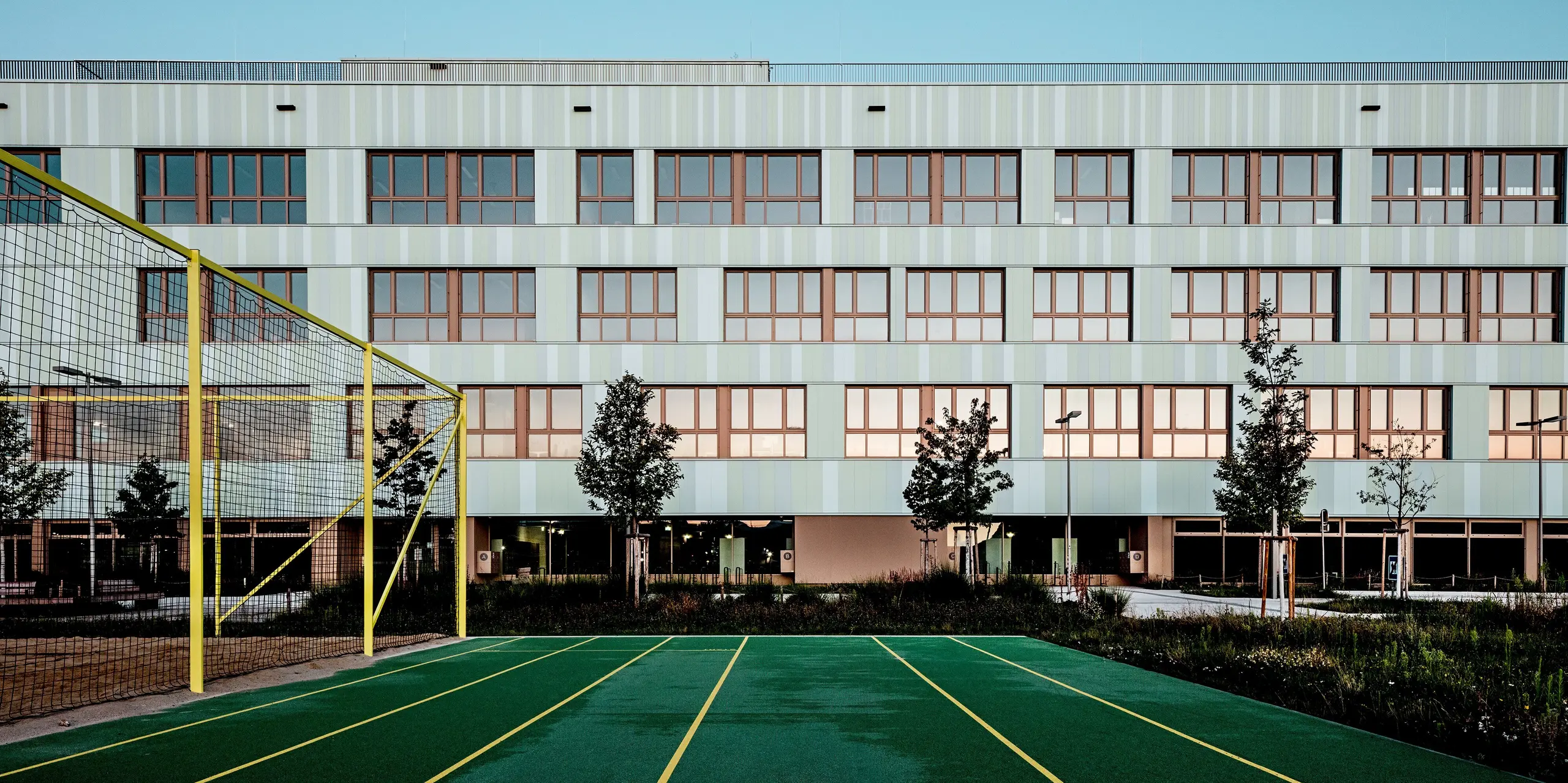 Das Bild zeigt die AHS Reininghaus in Graz mit ihrer modernen und ästhetischen Fassade aus PREFA Sidings in Sonderfarben. Die sanften Grüntöne der Fassade harmonieren perfekt mit den markanten braunen Fensterrahmen, die dem Gebäude einen eleganten Kontrast verleihen. Im Vordergrund ist ein Sportplatz mit einem grünen Laufband und gelben Markierungen zu sehen, was die Nutzung des Gebäudes für Bildung und Sport unterstreicht. Der Platz ist von schön angelegten Grünflächen und Bäumen umgeben, die das moderne Design der Architektur ergänzen. Die Verwendung von PREFA Sidings bietet nicht nur optische Schönheit, sondern auch herausragende Langlebigkeit und Widerstandsfähigkeit gegen Witterungseinflüsse. Die Kombination aus ansprechendem Design und funktionalen Elementen wie der Sporteinrichtung und den gepflegten Außenbereichen macht die AHS Reininghaus zu einem herausragenden Beispiel für zeitgenössische Architektur und nachhaltiges Design.