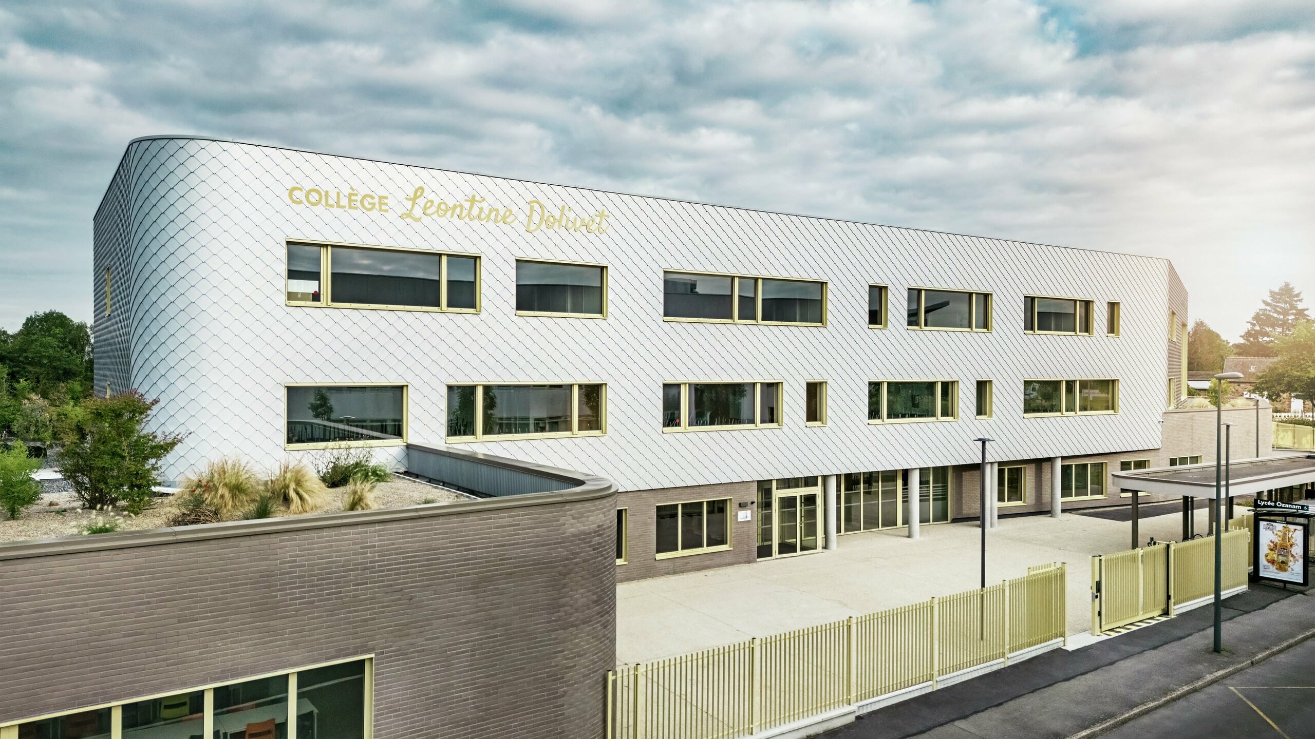 Frontansicht des Collège Léontine Dolivet in Cesson-Sévigné mit moderner Aluminiumfassade aus PREFA Wandrauten in Silbermetallic. Die changierende Oberfläche verleiht dem Gebäude Leichtigkeit, während klare Linien und große Fenster für Transparenz sorgen. Gelbe Fensterrahmen setzen lebendige Akzente. Im Vordergrund sind ein heller Vorplatz, ein Zaun und die angrenzende Straße sichtbar. Die langlebige PREFA Fassadenlösung kombiniert Design, Witterungsbeständigkeit und Nachhaltigkeit – ein architektonischer Blickfang mit eleganter Formensprache.