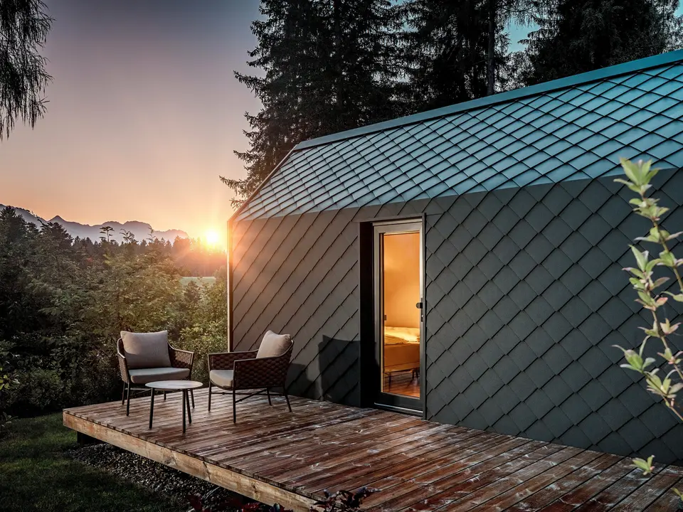 Ein minimalistisches Tiny House im Secret Koi Garden am Natterer See zeigt sich bei Sonnenuntergang mit vorgelagerter Holzterrasse und Blick in den Wald. Dach und Fassade sind mit kleinteiligen PREFA Dach‑ und Wandrauten aus Aluminium in P.10 Anthrazit ausgeführt, deren strukturierte Oberfläche das warme Licht eindrucksvoll reflektiert. Das langlebige, witterungsbeständige PREFA Aluminium schützt die Architektur zuverlässig und schafft einen markanten Kontrast zu Holz, Natur und sanfter Beleuchtung.