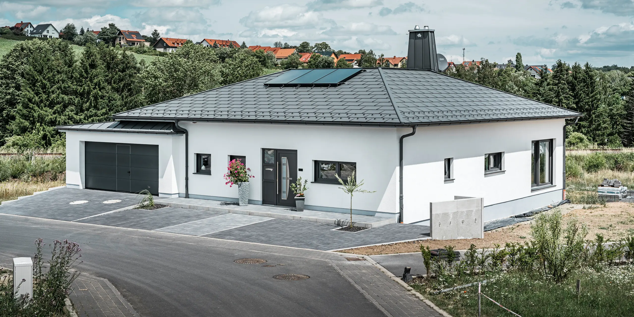 Elegant enfamiljshus i Ilmenau, Thüringen, med ett högkvalitativt PREFA-tak i färgen P.10 mörkgrå, utrustat med den robusta Takplatta R.16. Garaget harmonierar i färg och stil tack vare användningen av PREFALZ. Fastighetens blickfång är det funktionellt utformade atriumet och skorstenen dekorerad med PREFA-tjuren, som understryker PREFAs moderna design och innovativa takteknik. Solpanelerna smälter sömlöst in i husets hållbara koncept. Det genomtänkta helhetskonceptet för bungalowen kompletteras av PREFA:s takrännesystemet, som också är mörkgrått.
