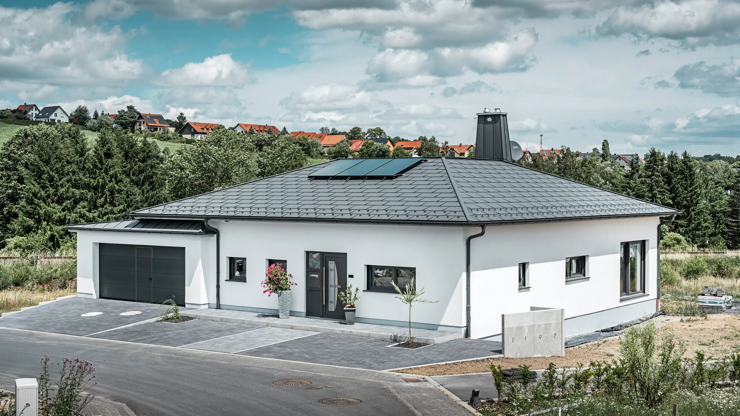 Elegant enfamiljshus i Ilmenau, Thüringen, med ett högkvalitativt PREFA-tak i färgen P.10 mörkgrå, utrustat med den robusta Takplatta R.16. Garaget harmonierar i färg och stil tack vare användningen av PREFALZ. Fastighetens blickfång är det funktionellt utformade atriumet och skorstenen dekorerad med PREFA-tjuren, som understryker PREFAs moderna design och innovativa takteknik. Solpanelerna smälter sömlöst in i husets hållbara koncept. Det genomtänkta helhetskonceptet för bungalowen kompletteras av PREFA:s takrännesystemet, som också är mörkgrått.