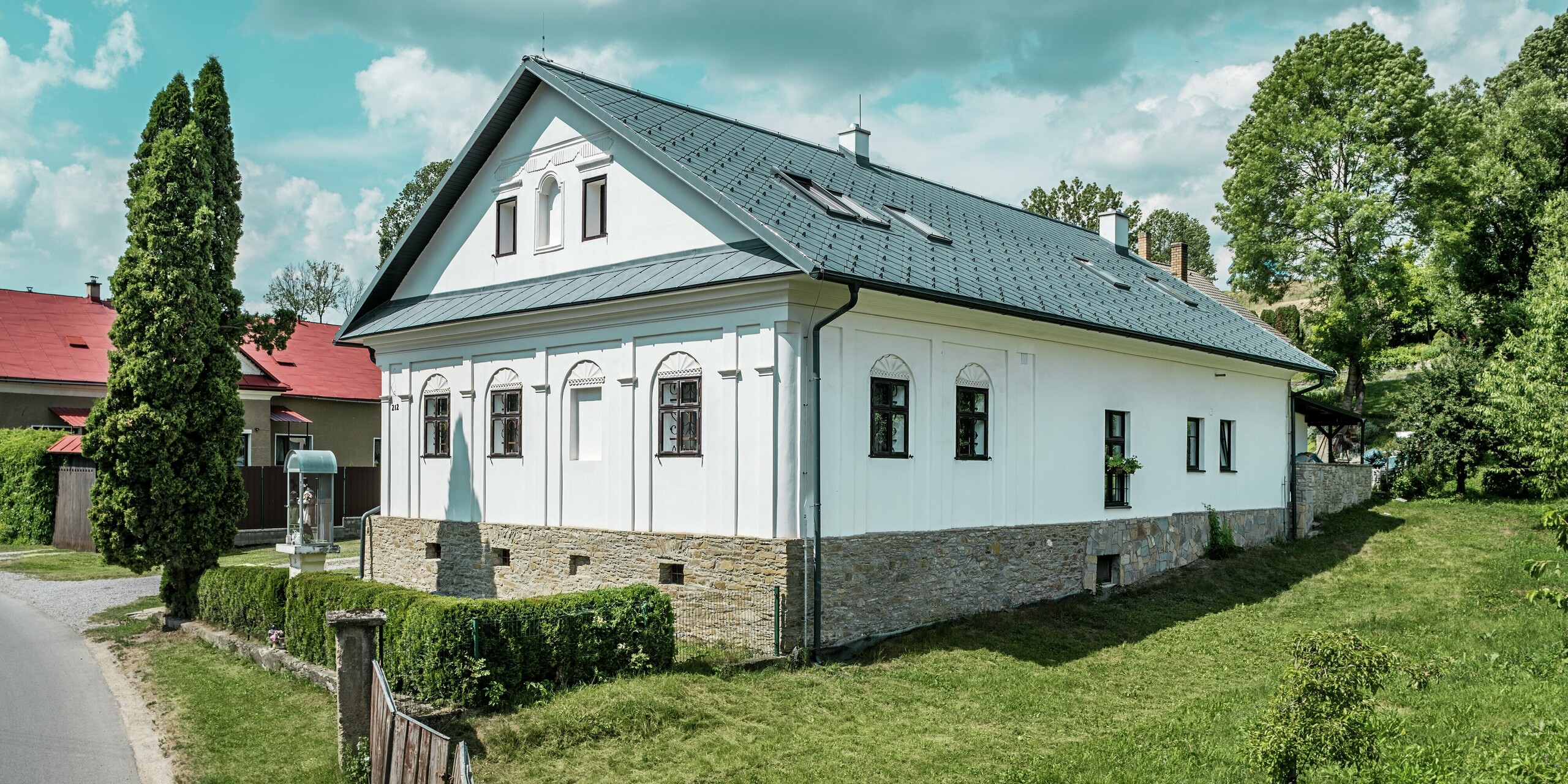 Das Bild von der Straße zeigt die Front und Seite des sanierten Handwerkerhauses in Klin, Slowakei, mit dem modernen Dach aus PREFA Dachrauten 44 × 44 in P.10 Steingrau. Der Blick auf die Gebäudeseite zeigt das elegante Satteldach und die perfekt integrierten Dachfenster, die für ein helles, offenes Ambiente sorgen. Die Dachrauten aus Aluminium ergänzen die traditionelle weiße Fassade und das Natursteinmauerwerk, was dem Haus einen zeitgemäßen und gleichzeitig klassischen Charakter verleiht. Mit ihren Vorteilen wie Langlebigkeit, Leichtigkeit und Korrosionsbeständigkeit ist die Dachlösung nicht nur ästhetisch ansprechend, sondern auch besonders funktional und nachhaltig.