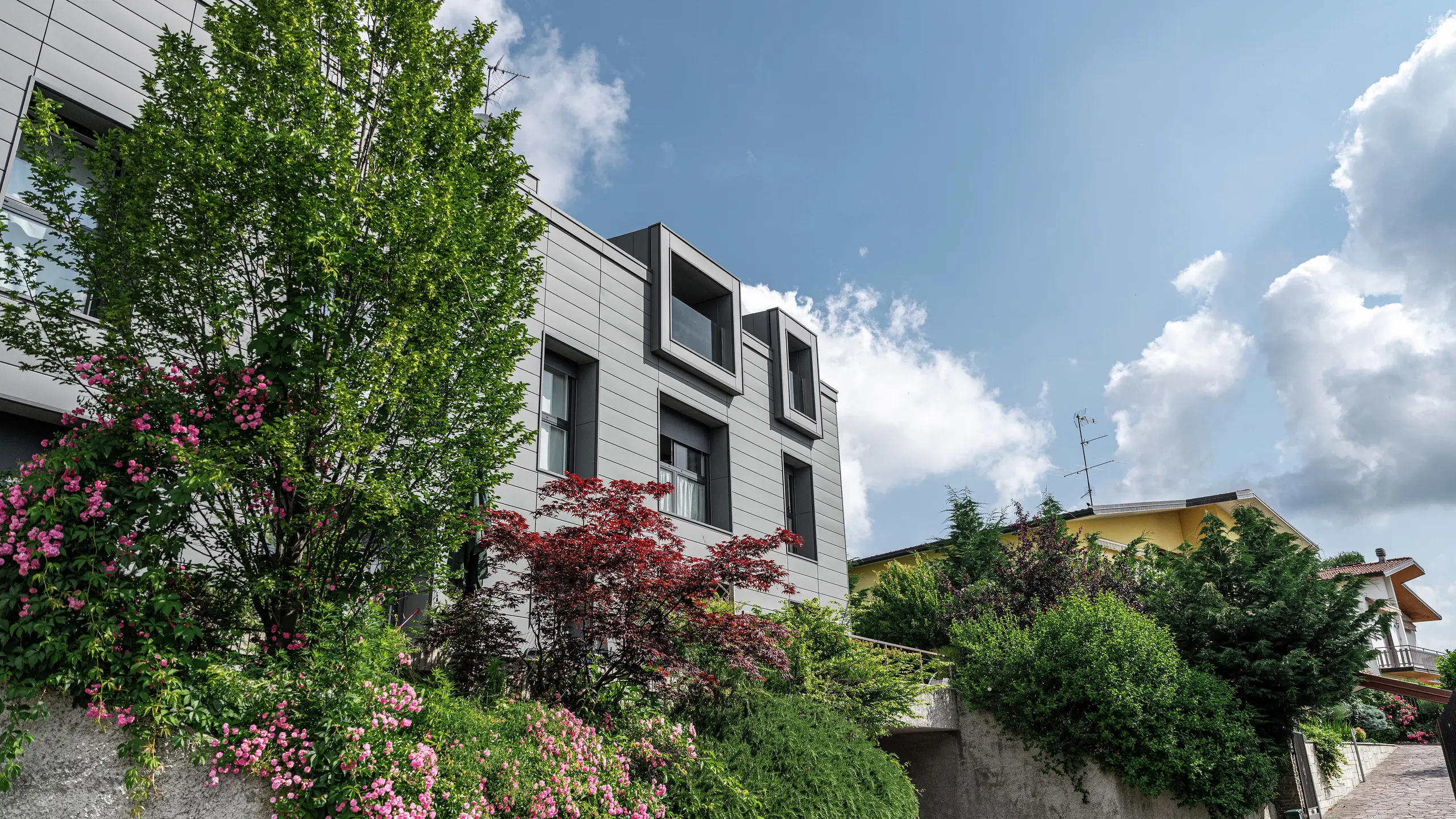 Modernt bostadsområde i Faloppio, Italien, med stilfull fasadpanel av PREFA ”Sidings”. De utskjutande fönstren är ett unikt arkitektoniskt blickfång, medan de omgivande växterna och blommorna understryker objektets naturliga skönhet. Den hållbara aluminiumbeklädnaden från PREFA är en estetisk och funktionell lösning i alla väder.