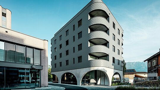 Dieses moderne Bernerhaus in Buchs besticht durch seine markant gerundete, dreieckige Gebäudeform und seine durchgängige Fassade aus PREFA Wandschindeln in der Farbe P.10 Hellgrau. Die schuppenartige Aluminiumoberfläche betont die weichen Kanten und tiefen Balkonrundungen, während der massive Betonsockel mit eleganten Arkaden einen spannenden Kontrast schafft. Vor alpiner Kulisse inmitten einer kleinen Einkaufsstraße wird die leichte, witterungsbeständige und langlebige PREFA-Aluminiumfassade zum architektonischen Eyecatcher: Sie ist präzise verarbeitet, wartungsarm und ideal für anspruchsvolle Rundungen.