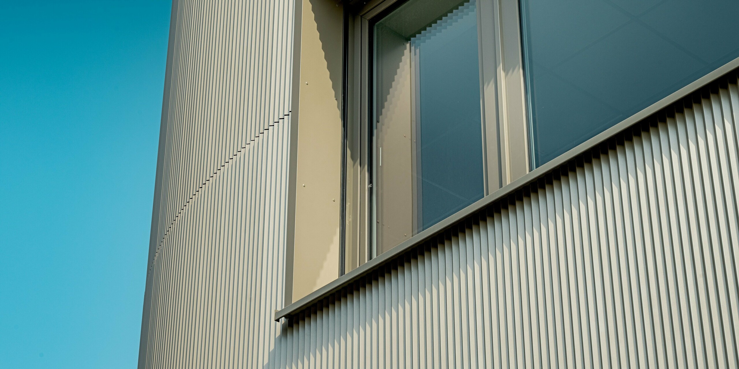 Das Bild zeigt ein Detail der Fassade des Festo-Gebäudes in Langenhagen, das mit PREFA Strangpressprofilen in Zackenform ausgestattet ist. Die vertikale Struktur der Profile erzeugt eine markante Textur, die zusammen mit den klaren Fensterrahmen eine moderne und dynamische Ästhetik bietet. Das sanfte Spiel von Licht und Schatten, das durch die geprägte Fassade entsteht, verstärkt die visuelle Wirkung des Gebäudes und sorgt für eine spannende Detailbetrachtung. Die robusten PREFA Materialien gewährleisten eine langlebige und pflegeleichte Oberfläche, die die architektonische Vision dieses Gebäudes über Jahre hinweg bewahrt.