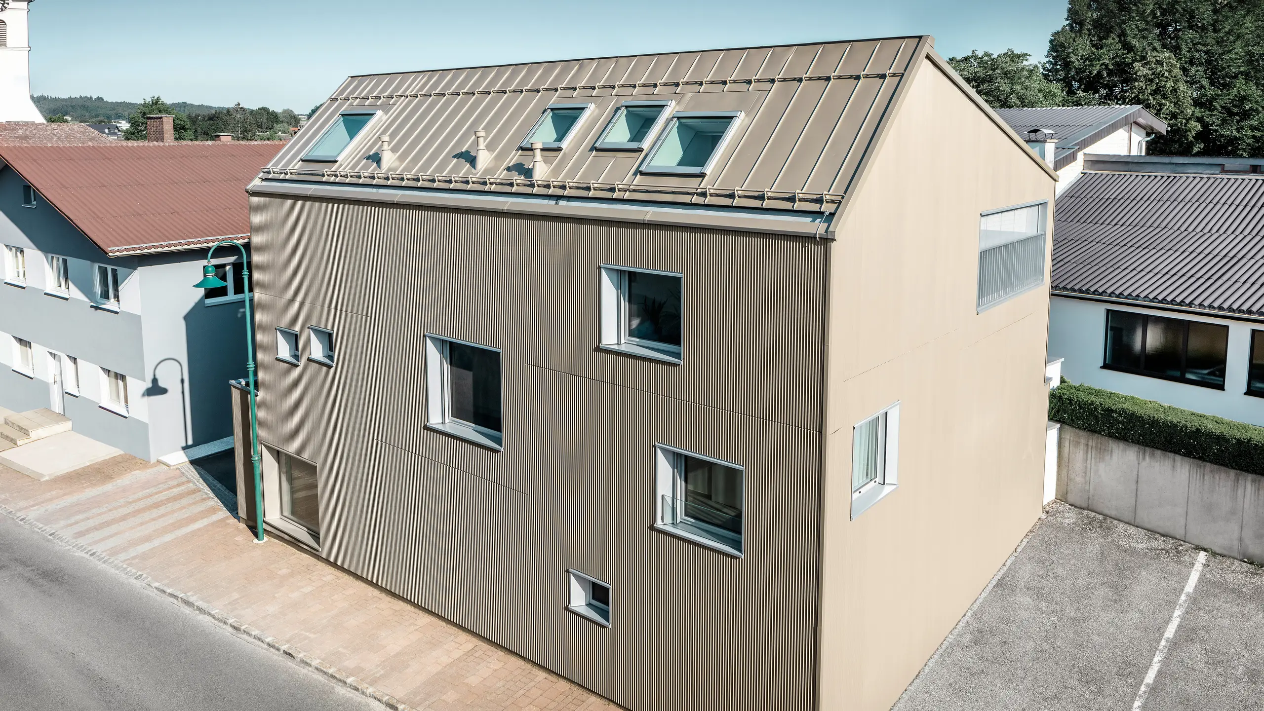 Fågelperspektiv på en villa i Frankenburg, Österrike, som sticker ut med sin PREFA-fasad i form av en tandad profil och ett PREFALZ-tak, båda i matchande bronsfärg. Fasaden har en markant struktur som fångar upp det naturliga ljuset och skapar ett levande skuggspel. Flera takfönster släpper in naturligt ljus och understryker den funktionella och estetiska integrationen av moderna arkitektoniska element. Huset smälter harmoniskt in i den typiska österrikiska bymiljön med sin moderna design och skapar samtidigt en kontrastrik accent mot de omgivande traditionella byggnaderna.