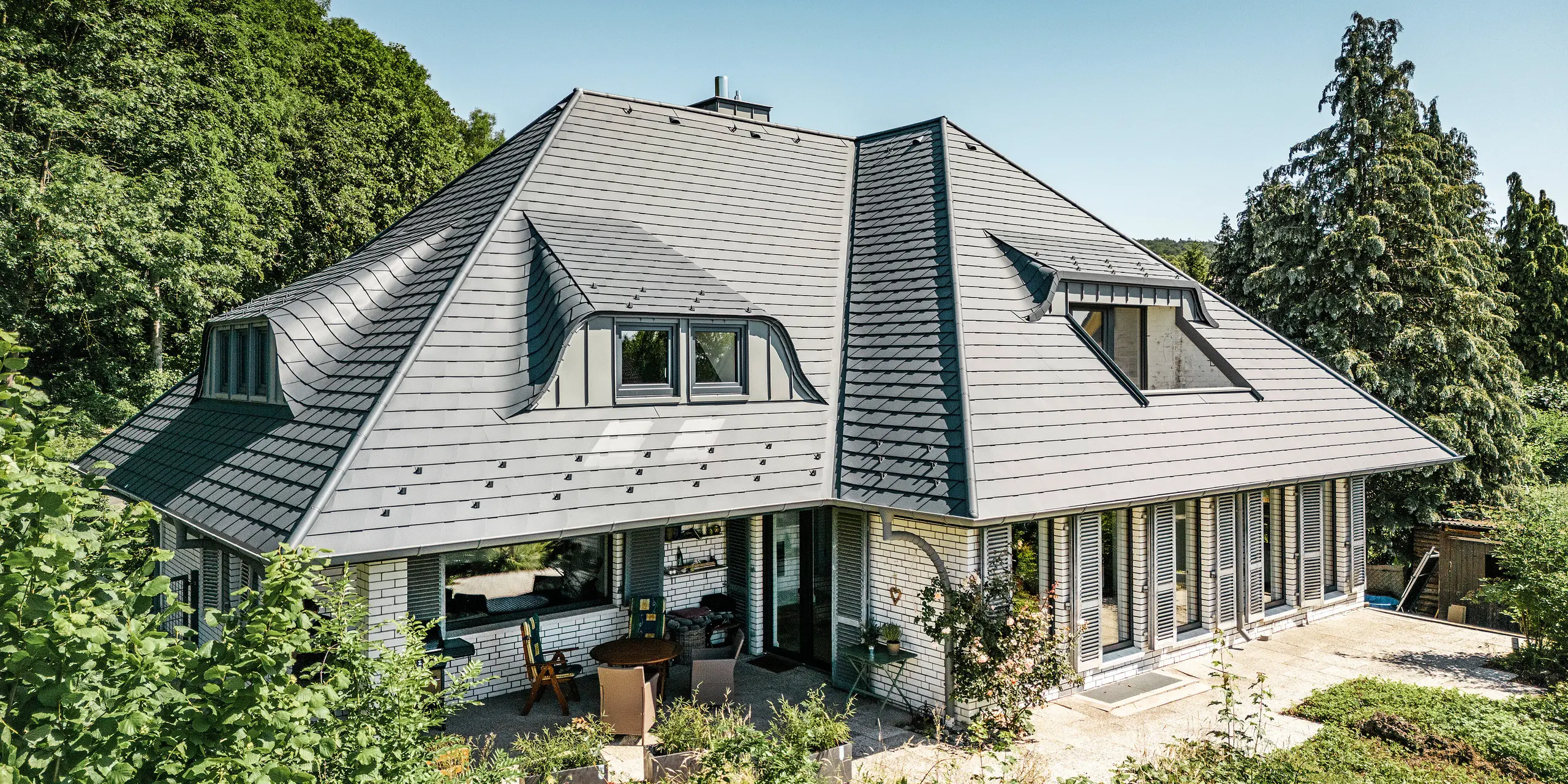 Modernt enfamiljshus i Einbeck, Tyskland, med PREFA takspån i P.10 antracit. De hållbara PREFA takspån av aluminium är kända för sin kvalitet och långa livslängd. Takkonstruktionen på byggnaden framhäver den individuella arkitekturen med takkupor, som lyfts fram av PREFA takspånens flexibilitet. Takavvattningen i form av PREFA-avloppsrör respektive hängränna är också i antracit. Runt enfamiljshuset finns en generös och välskött trädgård samt en terrass. Bakom huset ligger en skog som tillsammans med den blå himlen skapar en naturlig atmosfär.