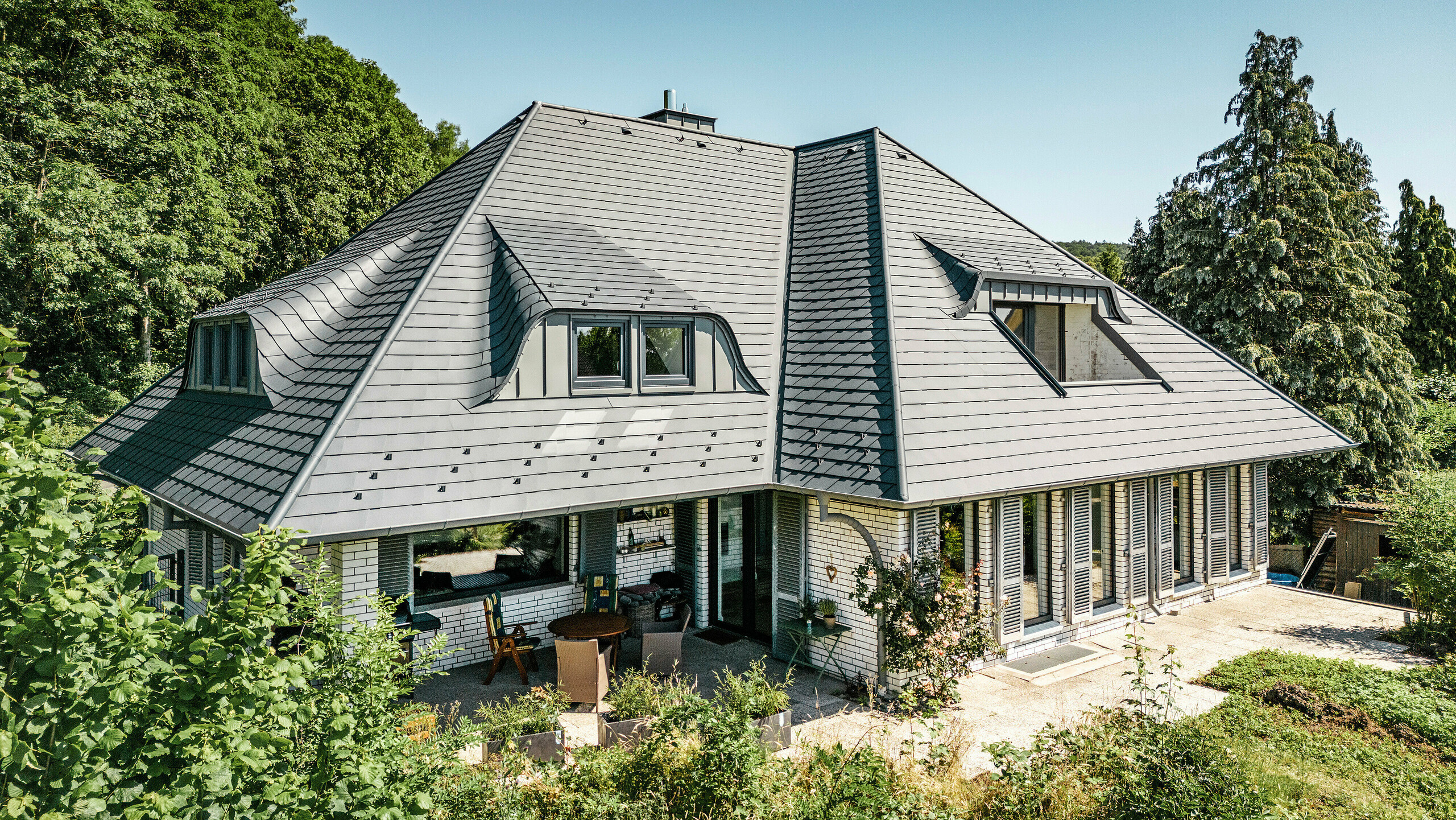 Modernt enfamiljshus i Einbeck, Tyskland, med PREFA takspån i P.10 antracit. De hållbara PREFA takspån av aluminium är kända för sin kvalitet och långa livslängd. Takkonstruktionen på byggnaden framhäver den individuella arkitekturen med takkupor, som lyfts fram av PREFA takspånens flexibilitet. Takavvattningen i form av PREFA-avloppsrör respektive hängränna är också i antracit. Runt enfamiljshuset finns en generös och välskött trädgård samt en terrass. Bakom huset ligger en skog som tillsammans med den blå himlen skapar en naturlig atmosfär.