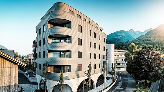 Schrägansicht des Bernerhauses in Buchs mit markanter, dreieckiger Kubatur und weich gerundeten Ecken. Die durchgehende Fassade aus PREFA Wandschindeln in P.10 Hellgrau umhüllt das Gebäude wie eine schuppenartige Aluminiumhaut und betont die fließenden Balkonbänder. Über dem massiven Betonsockel mit eleganten Arkaden wirkt die leichte, witterungsbeständige PREFA Aluminiumfassade besonders ausdrucksstark. Präzise verlegt, langlebig und wartungsarm fügt sie sich harmonisch in die alpine Umgebung ein und setzt ein starkes architektonisches Statement.