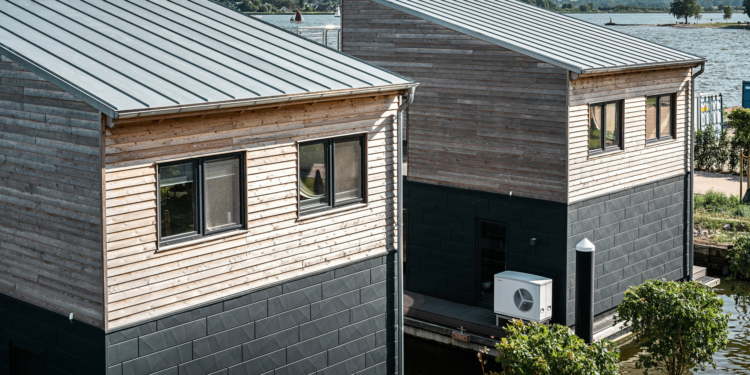 Drohnenaufnahme von zwei Wasserhäusern im Pionierhafen Schleswig, die eine attraktive Kombination aus naturbelassenen Holzoberflächen und dunklen PREFA Siding.X Aluminiumpaneelen in P.10 Anthrazit aufweisen. Die Architektur integriert moderne Haustechnik, wie die sichtbare Wärmepumpe, die für eine umweltfreundliche Klimaregulierung sorgt. Die Häuser sind durch ihre moderne Bauweise und robuste Materialwahl ideal an das Leben am Wasser angepasst, wodurch sie eine alternative Wohnoption darstellen.