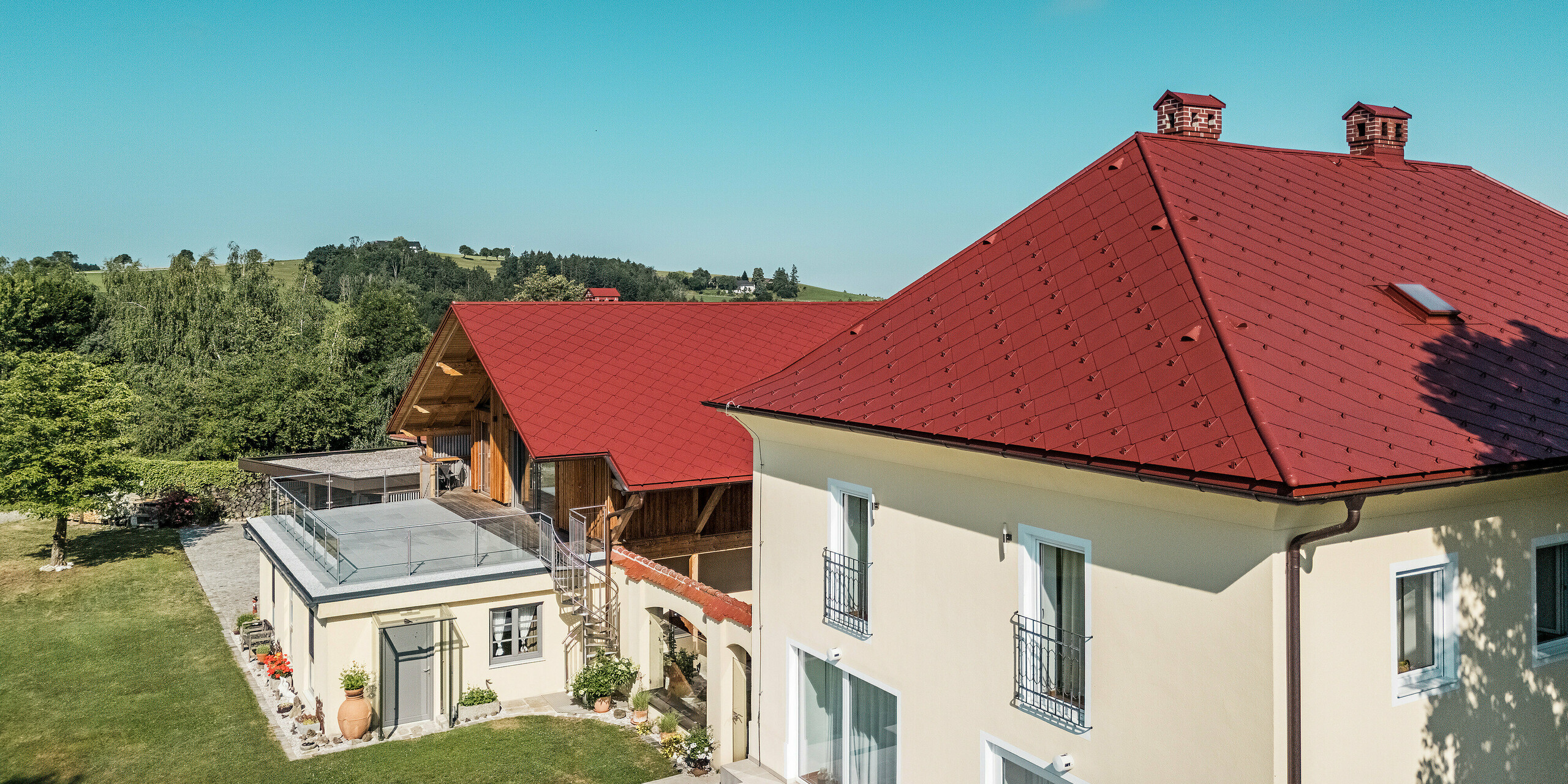 Das Bild zeigt die sanierten Einfamilienhäuser in Aschach an der Steyr aus der Luft. Das Walmdach und das Satteldach wurden mit PREFA Dachrauten 44 x 44 in der Farbe P.10 Oxydrot neu eingedeckt. Die roten Dächer setzen auffällige Akzente in der ländlichen Umgebung. 