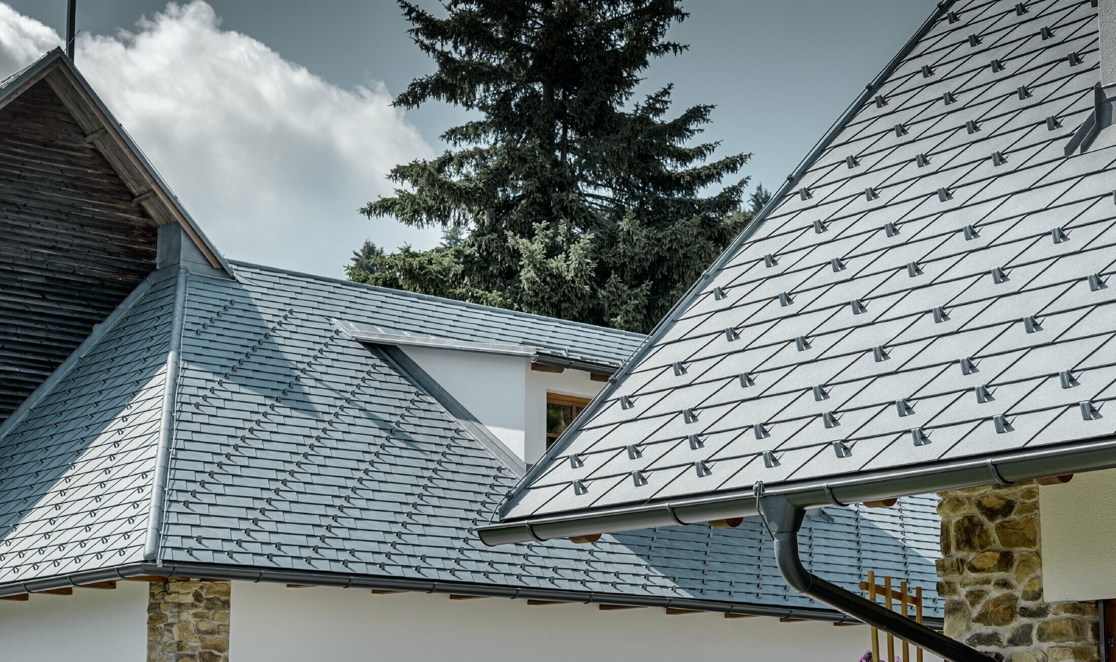 Detalj av aluminiumbeläggningen från PREFA - Takshingel i stengrått med takrännor i antracitgrå aluminium från PREFA. I bakgrunden ser en takkupa med stående fals. Fasaden är vit med inbyggda stenkomponenter.