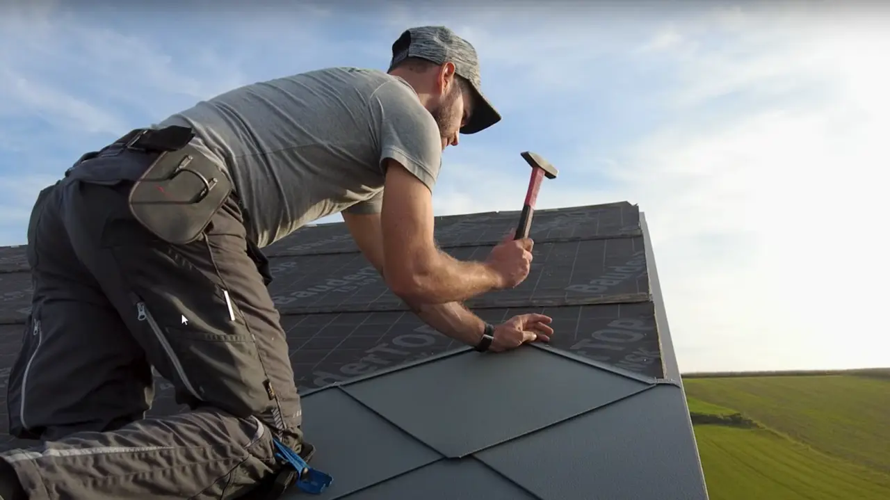 Plåtslagare i aktion: Med en hammare lägger ägaren till företaget „Der Blechmann“ noggrant PREFA takplattor 44 × 44. Precisionen vid installationen syns på de redan lagda plattorna längst ner i bilden. Mannen bär en mössa och knäböjer med ryggen mot kameran på taket. I bakgrunden syns den klara himlen och ett grönt landskap. Den harmoniska bakgrunden understryker bildens symbolik: hållbart och långvarigt hantverk med högkvalitativa PREFA-aluminiumprodukter.