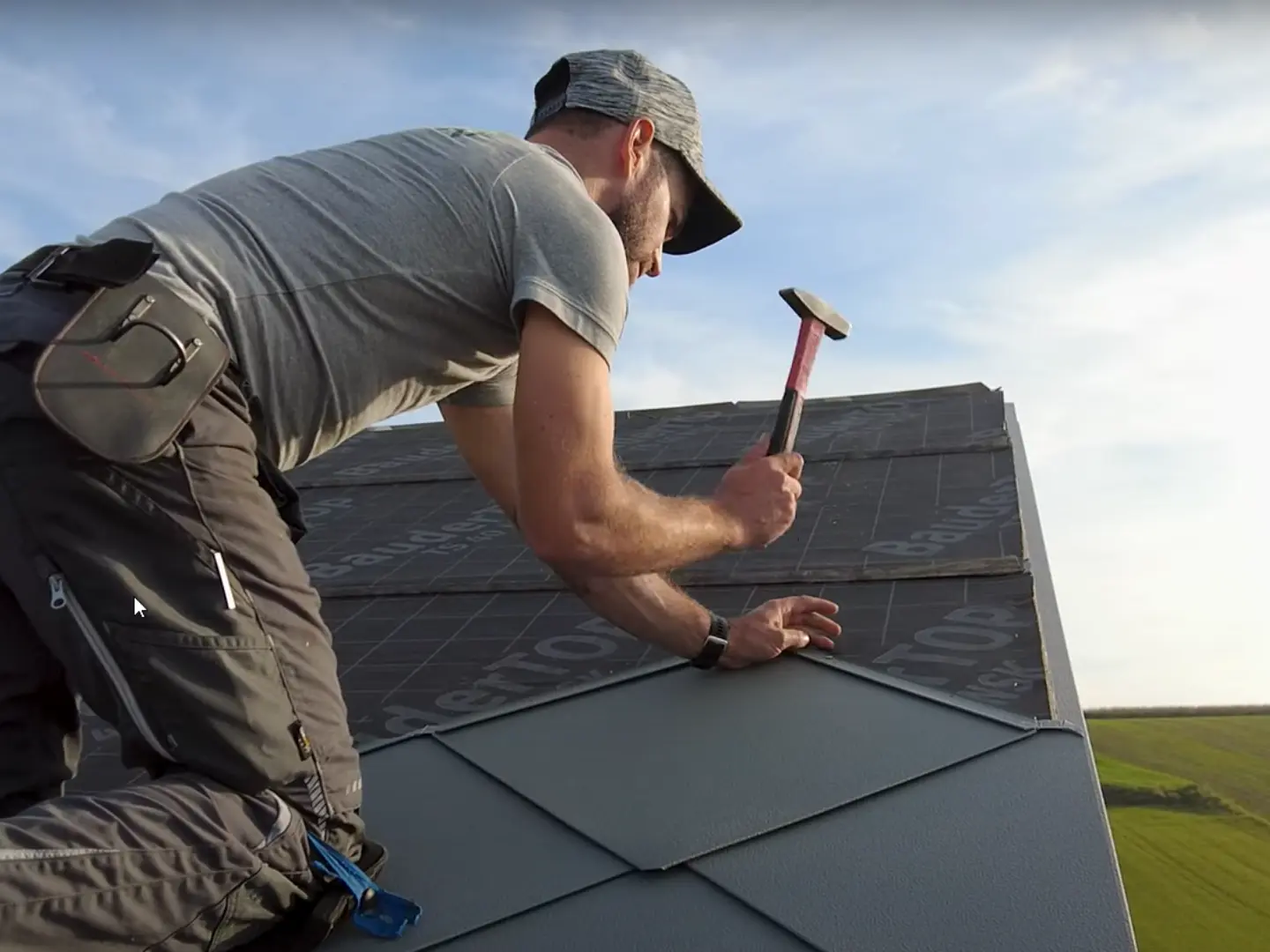 Plåtslagare i aktion: Med en hammare lägger ägaren till företaget „Der Blechmann“ noggrant PREFA takplattor 44 × 44. Precisionen vid installationen syns på de redan lagda plattorna längst ner i bilden. Mannen bär en mössa och knäböjer med ryggen mot kameran på taket. I bakgrunden syns den klara himlen och ett grönt landskap. Den harmoniska bakgrunden understryker bildens symbolik: hållbart och långvarigt hantverk med högkvalitativa PREFA-aluminiumprodukter.