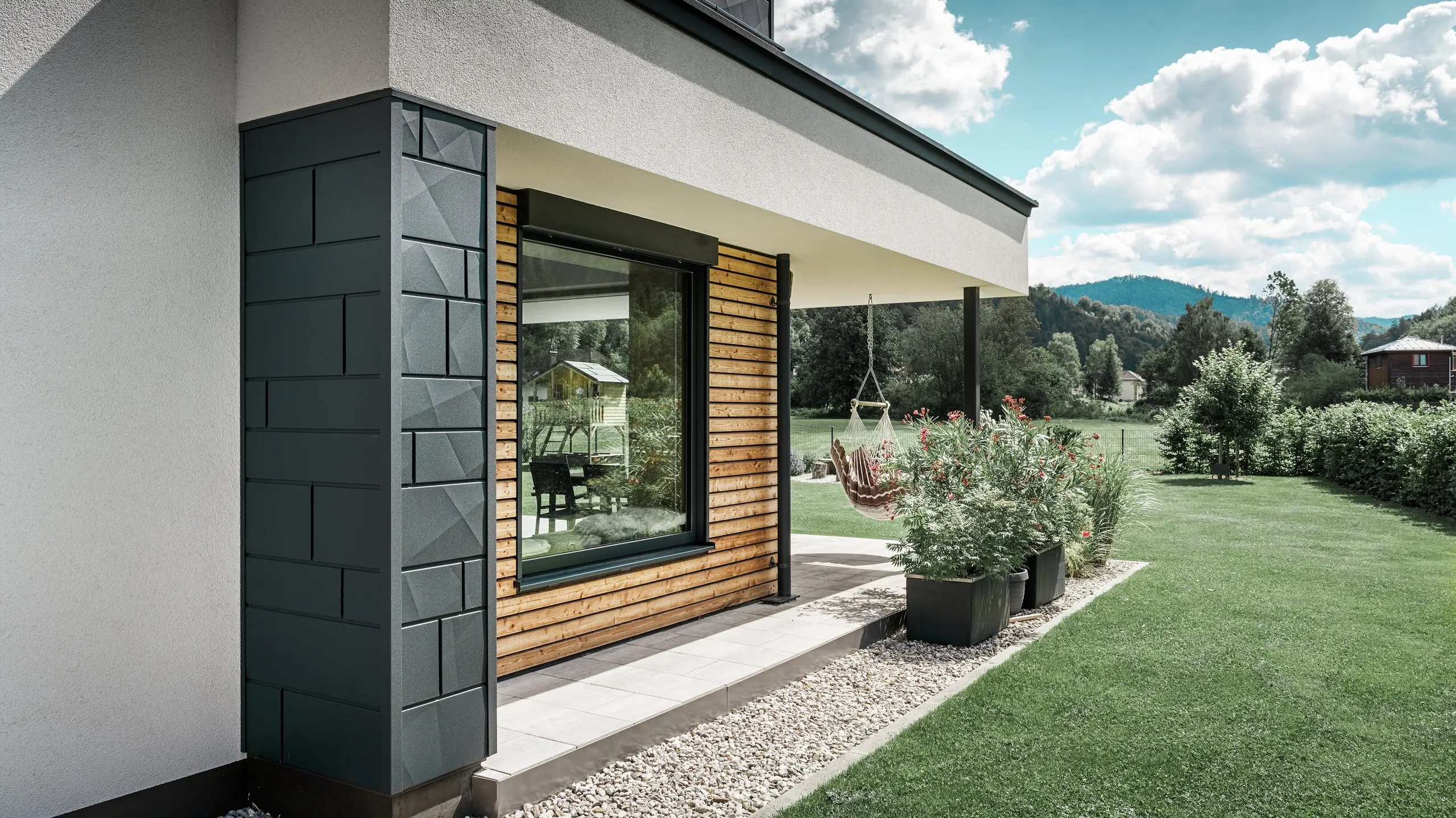 Partiell vy av ett nybyggt enfamiljshus. Här har olika material använts på fasaden. Den vitputsade fasaden framhävs av PREFA aluminium Fasadpanel.X i antracit och horisontella träpaneler, som omger ett stort fönster. På terrassen hänger en hängande stol.