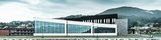 In der Abbildung ist das Healthcare Center in Oystese vom Wasser aus zu sehen. Diese spiegelt sich im Wasser des Hardangerfjord und wurde mit der Wandraute 44x44 in Naturblank eingedeckt. Der Himmel dahinter ist Wolkenbedeckt.