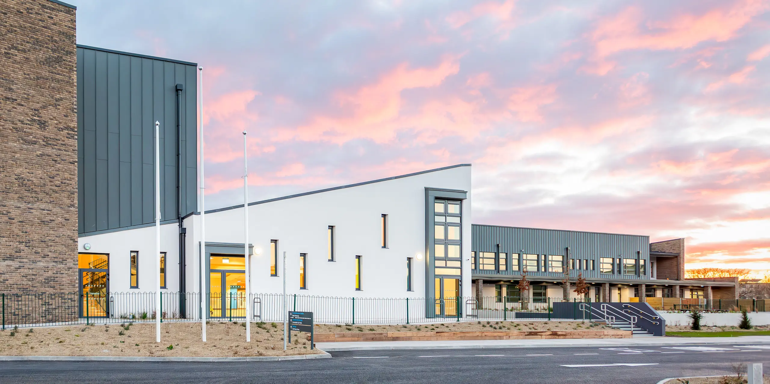 Das moderne Schulgebäude der Scoil Mhuire Naofa in Carrigtwohill, Irland, präsentiert sich mit einer markanten Aluminium-Fassade aus PREFALZ in P.10 Hellgrau. Die langlebige und witterungsbeständige Stehfalzdeckung umfasst rund 800 m² und verbindet Funktionalität mit zeitloser Ästhetik. Vor der Kulisse eines malerischen Sonnenuntergangs kommt die hochwertige Architektur besonders gut zur Geltung.