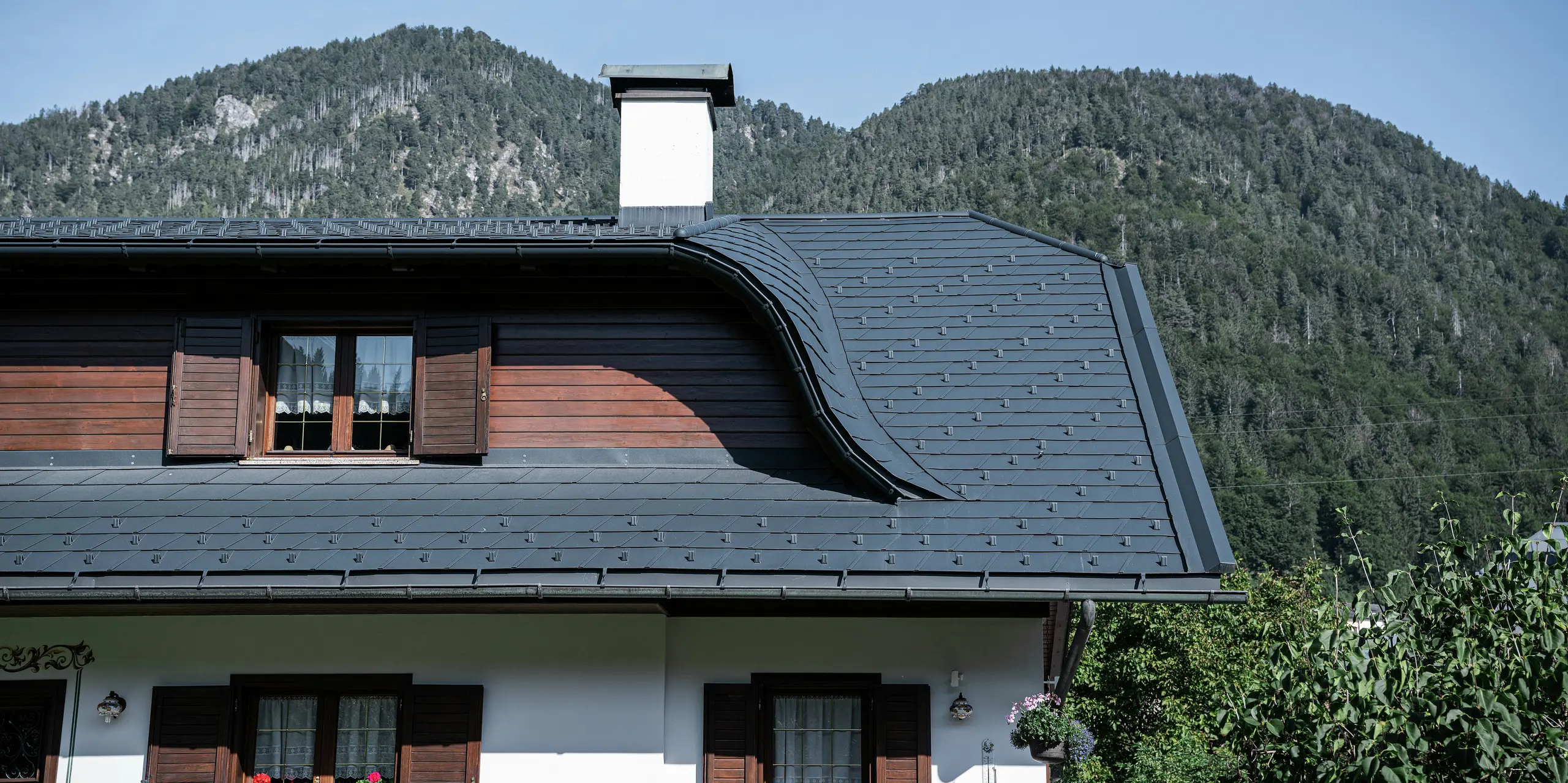 Nahaufnahme des Daches eines Einfamilienhauses in Tarvis, Italien, nach der Sanierung mit PREFA Dachschindeln P.10 anthrazit. Die elegante Hechtgaube verleiht dem Dach seine geschwungene Form und unterstreicht den traditionellen Stil des Hauses. Die langlebige Aluminium-Dacheindeckung bietet optimalen Schutz vor extremen Witterungseinflüssen. In Kombination mit der weißen Fassade und den Holzelementen entsteht ein harmonisches, alpines Gesamtbild, das sich perfekt in die Berglandschaft einfügt.