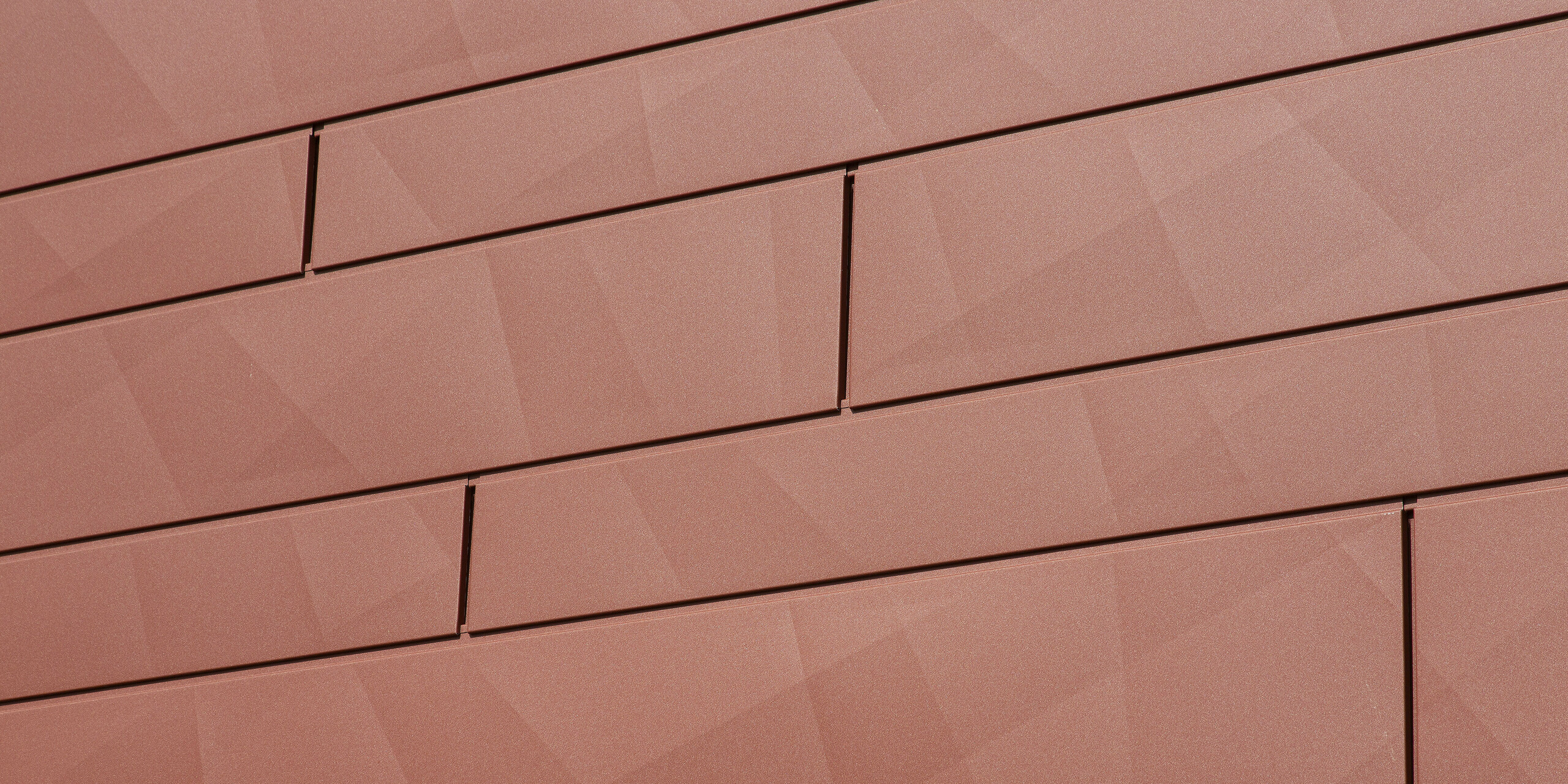 Detailaufnahme der PREFA Fassadenpaneele FX.12 in Oxydrot: Die prägnante Kantung erzeugt eine spannende geometrische Struktur, die das Licht je nach Tageszeit anders reflektiert. Die hochwertige Pulverbeschichtung in P.10 Qualität sorgt für dauerhafte Farbbrillanz und höchste Widerstandsfähigkeit – ideal für langlebige, wartungsfreie Fassadensanierungen.