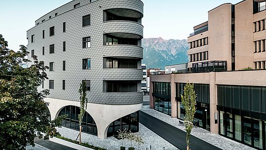 Ansicht des Bernerhauses in Buchs mit markanter, dreieckiger Form und weich gerundeten Balkonen vor alpiner Bergkulisse. Die durchgängige Fassade aus PREFA Wandschindeln in P.10 Hellgrau umhüllt das Gebäude wie eine schuppenartige Aluminiumhaut und erzeugt ein lebendiges Spiel aus Licht und Schatten. Über dem massiven Betonsockel mit eleganten Arkaden wirkt die leichte, witterungsbeständige PREFA Aluminiumfassade besonders präzise, langlebig und wartungsarm – ein architektonischer Eyecatcher im urbanen Umfeld.