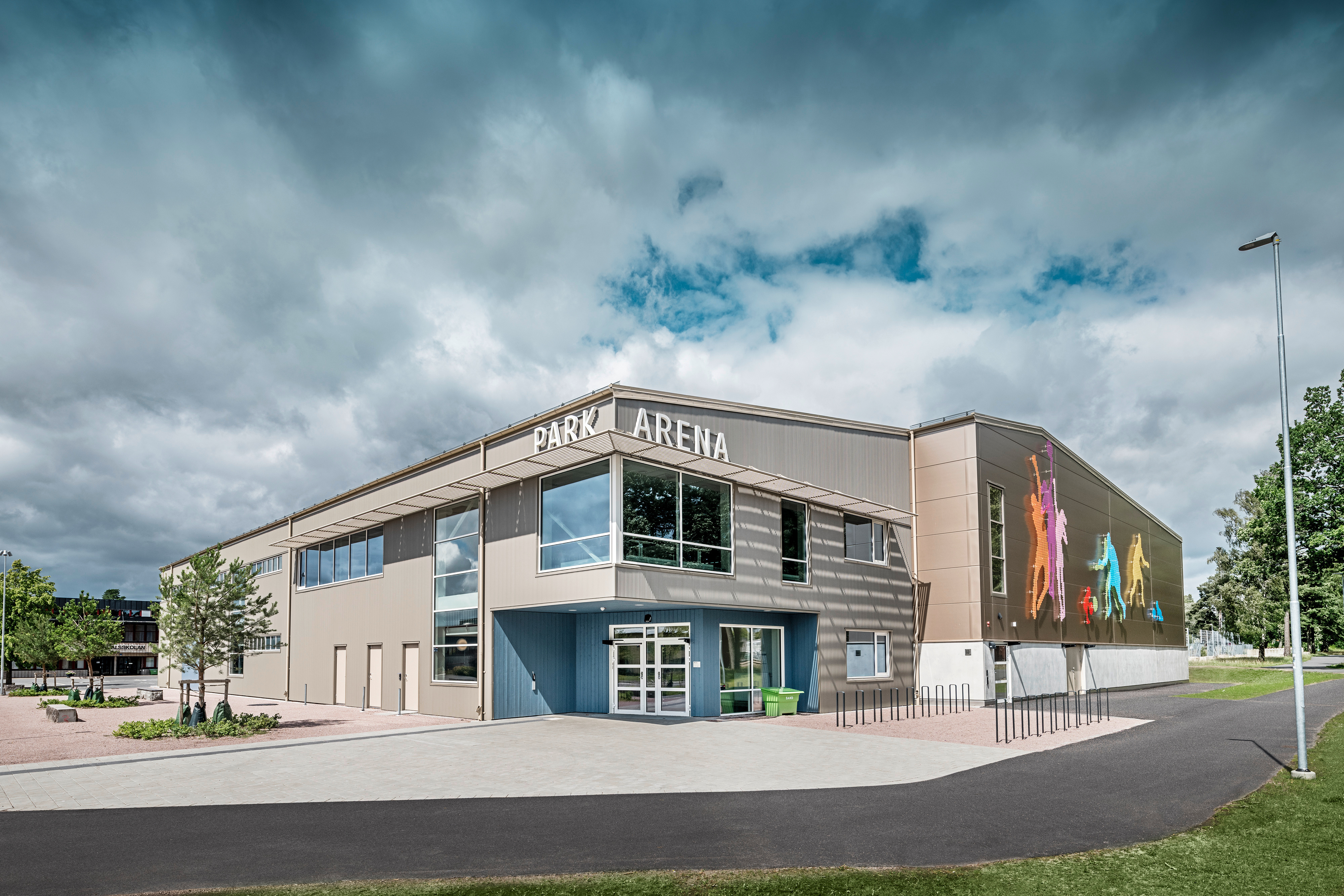 Frontansicht der Park Arena in Värnamo, Schweden, mit moderner PREFA Fassade aus Zackenprofilen in Bronze. Die Aluminiumfassade bietet eine langlebige und wetterbeständige Lösung, die durch markante Licht- und Schatteneffekte hervorsticht. Ein künstlerisch gestaltetes Wandbild auf der rechten Seite zeigt farbenfrohe sportliche Figuren, die den dynamischen Charakter der Sporthalle betonen. Große Fensterflächen lassen viel natürliches Licht in das Gebäude und verstärken das moderne Design der Arena.